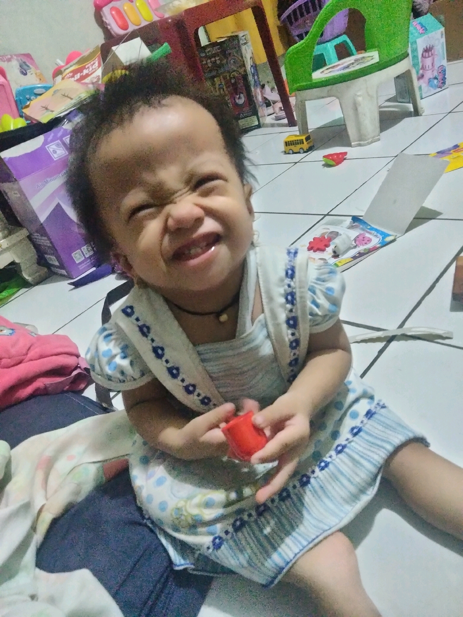 In Suka Sari, Indonesia, a young child with dark, curly hair sits on a tiled floor in a home. The child, wearing a light-colored dress with blue polka dots and a blue and white patterned trim, is holding a small red object. The child's face is contorted in a playful grimace, with eyes squeezed shut and mouth open, revealing teeth. The background reveals a domestic setting with various toys scattered around, including a yellow toy bus and a green plastic chair. A purple box with a graphic design is visible on the left, and a pink blanket or clothing item is on the child's lap. The lighting is natural, suggesting daytime.