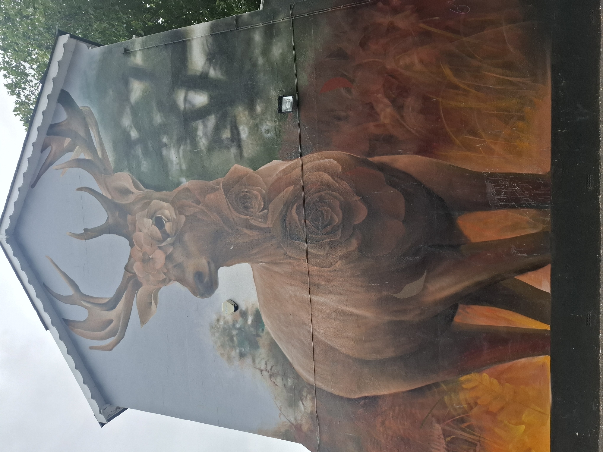 The image is a full shot of a mural painted on the side of a building in Ashford, United Kingdom. The mural depicts a large, brown stag with impressive antlers. Its head is adorned with a floral crown made of pale pink roses. The stag's body is also embellished with roses, one prominent rose on its chest and another further down its body. The background of the mural features a muted forest scene with the silhouettes of trees. The overall color palette is warm, with earthy tones and hints of orange and red in the foreground, suggesting foliage. The building itself has a grey, slightly textured wall. A white scalloped trim runs along the roofline. There is a light fixture and electrical wiring visible on the mural's surface. The mural is located on a residential or commercial building, as evidenced by the presence of a small, white security sensor on the wall. The bottom of the mural is bordered by a black plinth. The sky above is overcast and pale.