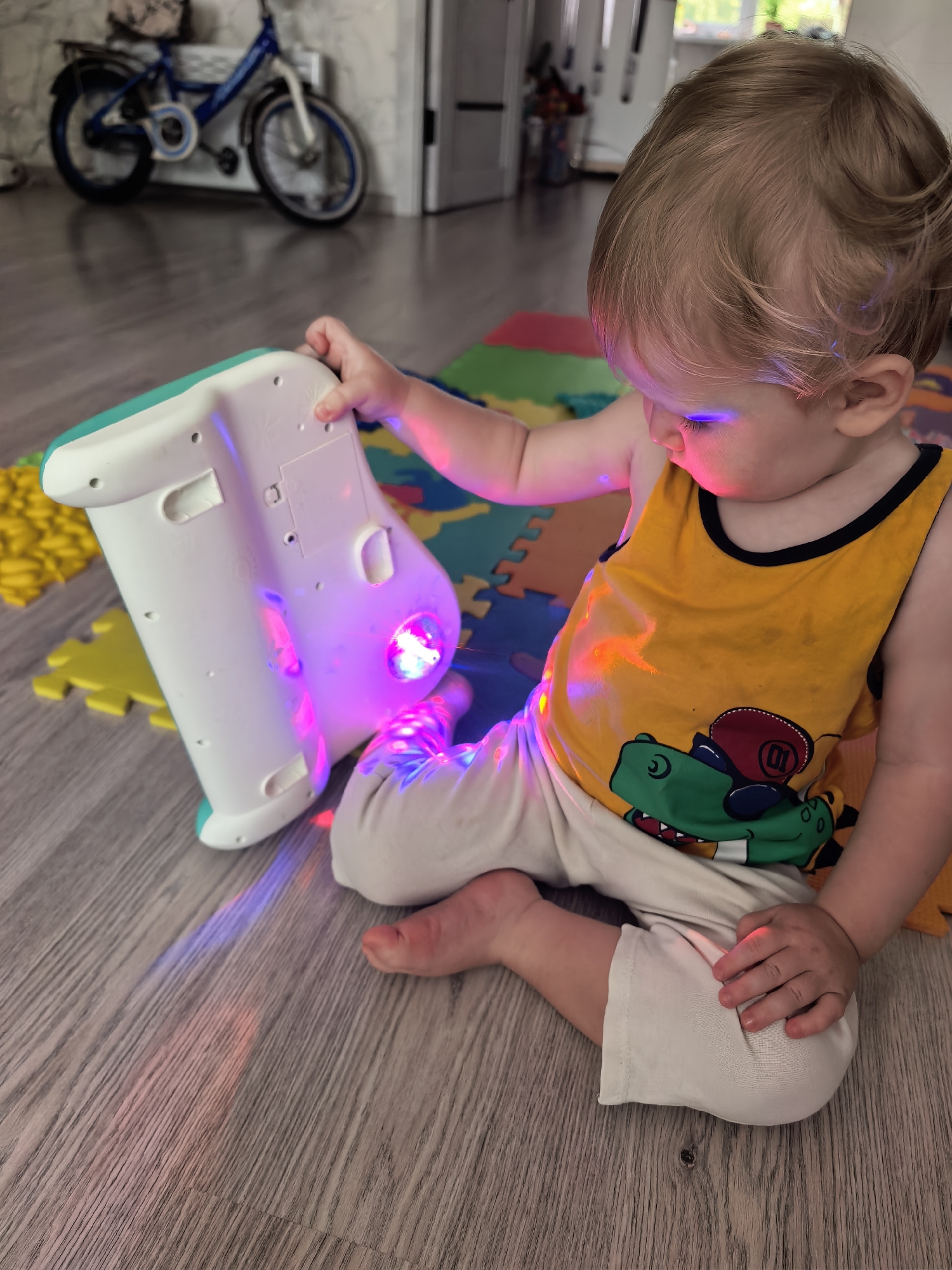 A light-skinned toddler with blonde hair sits on a wooden floor, playing with a white and teal electronic toy that projects colorful lights onto the floor and the toddler's face and clothing. The toy has a picture of a dinosaur on a yellow tank top worn by the toddler. The toddler is on a colorful puzzle mat. In the background, a blue bicycle with a basket is parked near a wall. The scene appears to be indoors, possibly in a home, and there is no specific indication of its location in Sterlitamak, Russia.