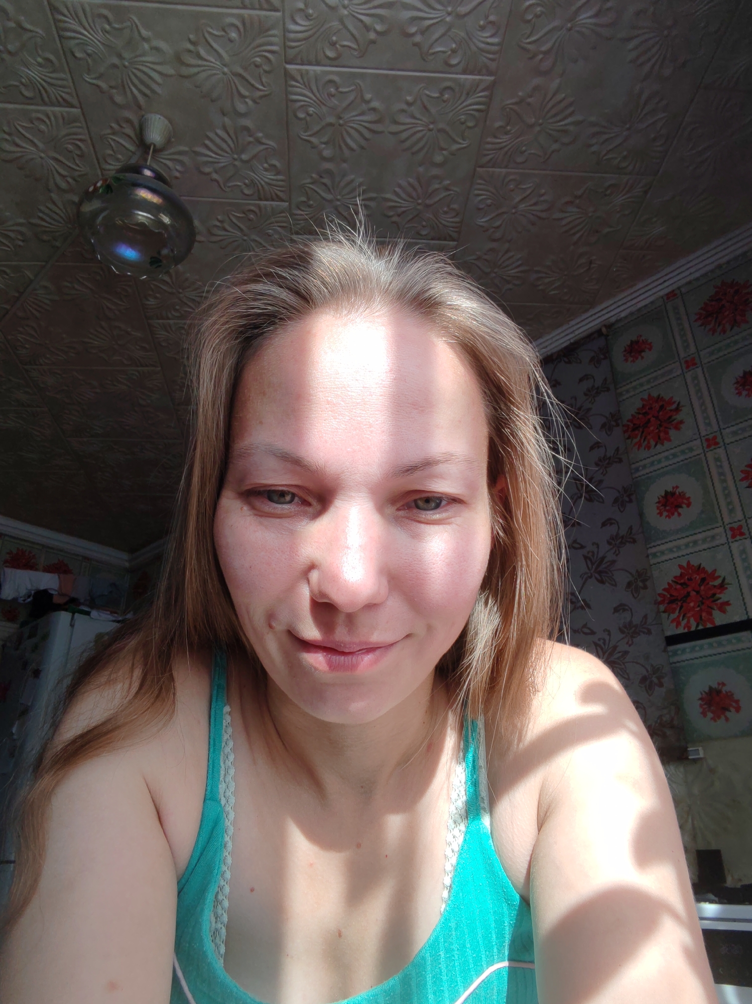 A mid-shot, head-and-shoulders photograph shows a woman with light brown hair and fair skin. She is looking slightly off-camera to her right, with a subtle smile. Sunlight illuminates her face from the left, casting shadows and highlighting her features. She is wearing a teal tank top with white lace trim. In the background, the ceiling is covered in textured panels, and the walls are adorned with floral wallpaper. A ceiling light fixture is visible in the upper left corner. The scene appears to be indoors, possibly in a residential setting. The image does not provide any specific information about the location within Чухлэм, Russia.