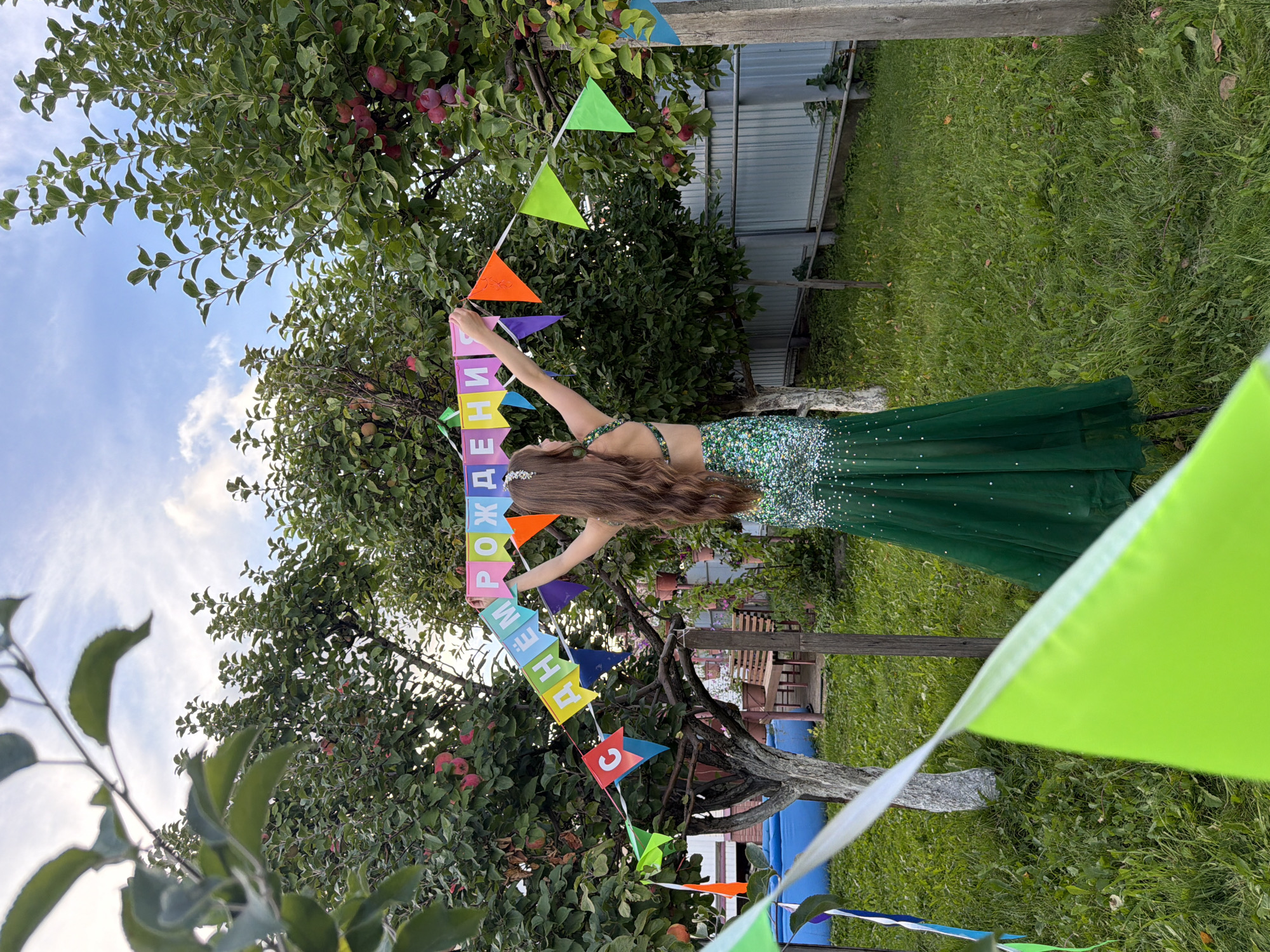 A young woman with long brown hair, wearing a sparkly green dress, stands in a backyard garden. She is holding up a string of colorful pennant flags that spell out "С ДНЕМ РОЖДЕНИЯ" (Happy Birthday in Russian). The garden appears to be in a residential area, with apple trees and a fence in the background. The sky is partly cloudy. The overall scene suggests a birthday celebration taking place outdoors. The location is identified as Chaikovsky, Russia.