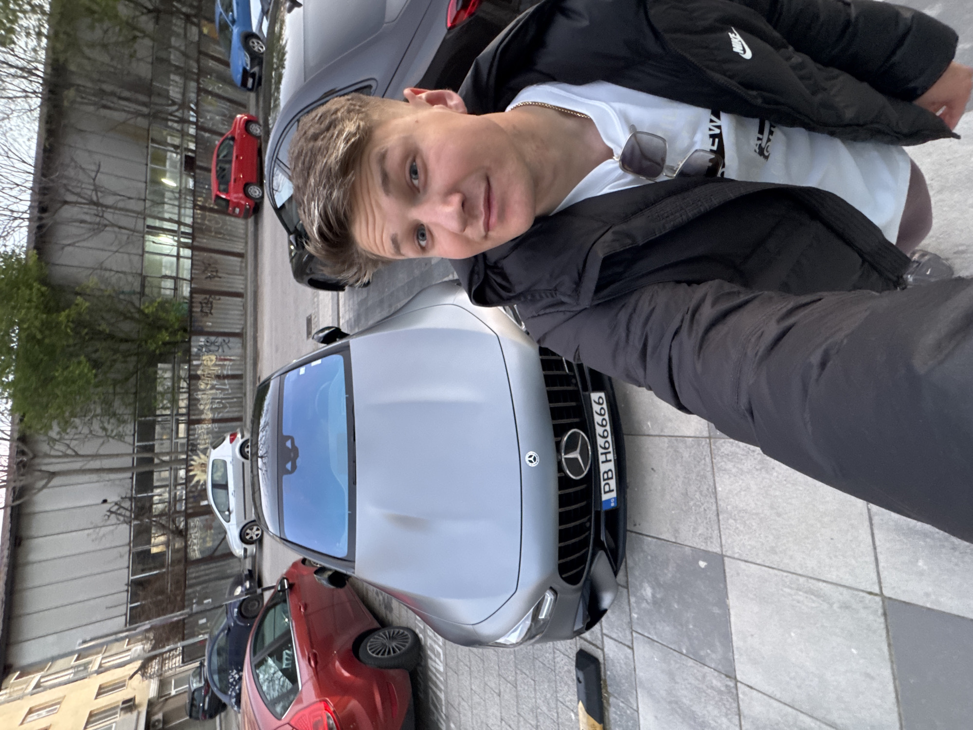 The image captures a person taking a selfie with a silver Mercedes-Benz with Bulgarian license plate "PB H6666" parked on a city street. The person is wearing a white T-shirt under a black Nike jacket and has a pair of sunglasses attached to their shirt. They have short, light-colored hair and blue eyes. Behind the Mercedes-Benz, there are several other parked cars of various colors (red, blue, white). In the background, there's a building, trees, and a metal structure that appears to be part of a fence or a scaffolding system. The setting is a city street in Plovdiv, Bulgaria, based on the Bulgarian license plate of the car.