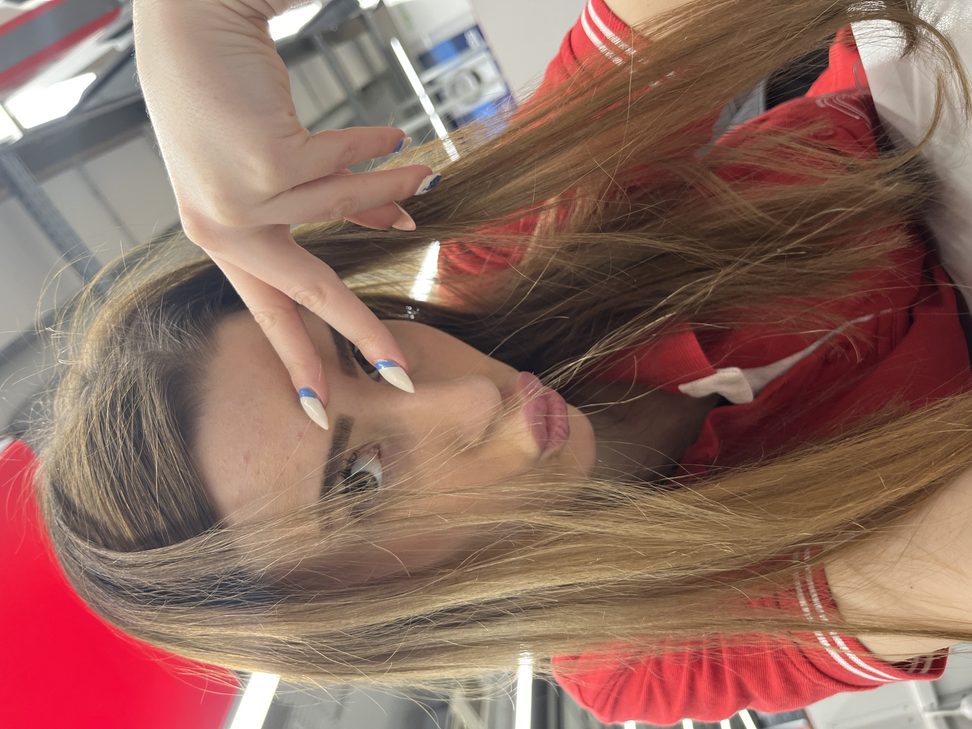A woman is photographed close-up, angled downward from her left side. She has fair skin, brown hair, and light-colored eyes. Her hand is raised near her face, displaying long, white nails with a blue design. She is wearing a red top with white stripes on the sleeves. The background is blurry, with hints of gray and white, possibly an indoor setting. The photograph's location is unknown, so I cannot confirm if it is in Sofia, Bulgaria.