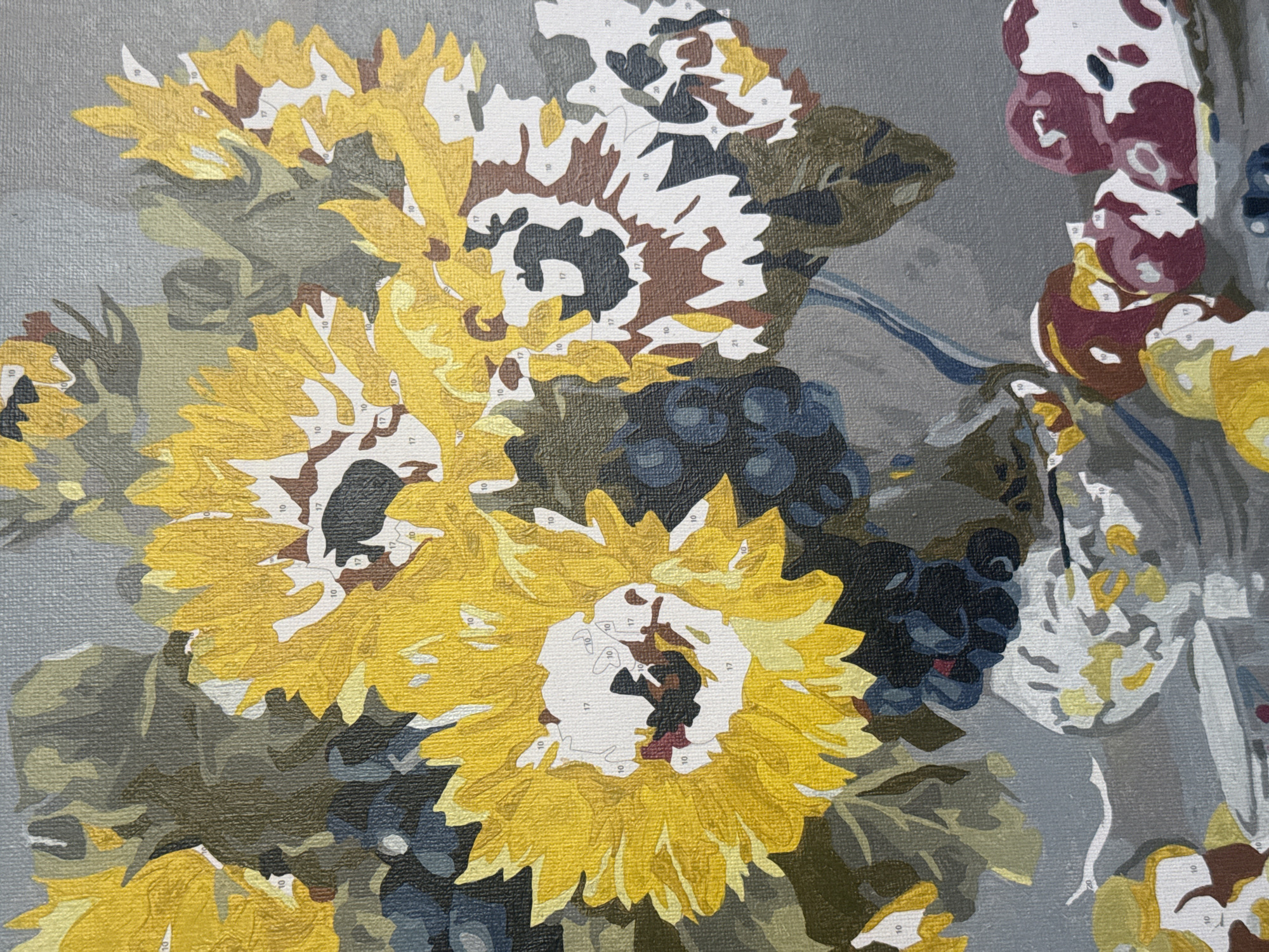 The image depicts a close-up of a paint-by-numbers canvas. The subject matter appears to be a still-life arrangement of sunflowers, grapes, and possibly other floral elements. The canvas is pre-printed with numbered outlines indicating which colors should be applied to each section. The background is a neutral gray tone. The paint has not yet been applied, as the numbers are clearly visible within each section. The lighting is even and diffuse.
The location in Sofia, Bulgaria is irrelevant as the painting is not a scene of the city, or located there.