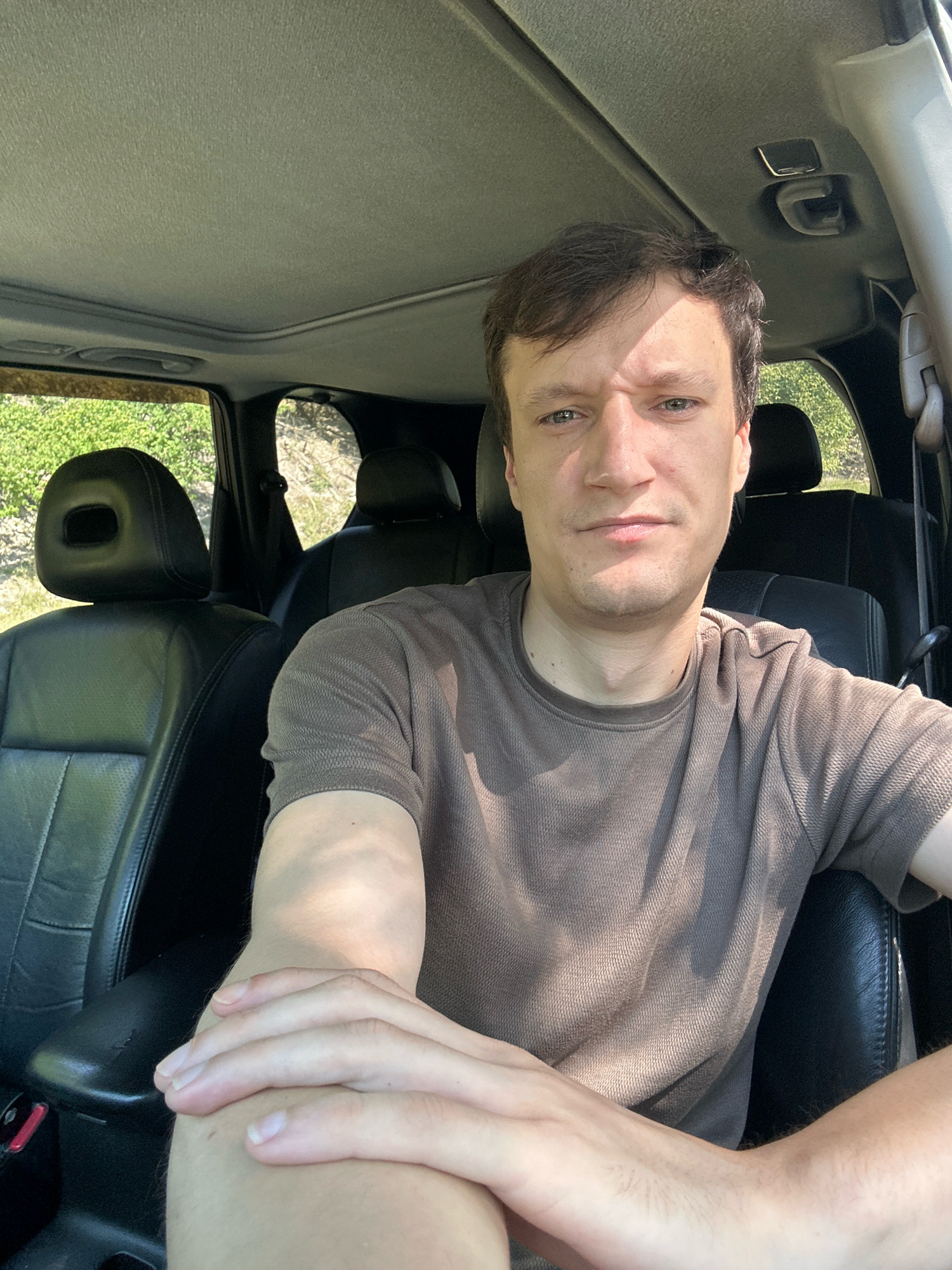 A man with brown hair is sitting in the driver's seat of a car. He is wearing a brown t-shirt. The car's interior is visible, including black leather seats and a sunroof. Outside the car, green foliage and some dry vegetation are visible. The scene appears to be outdoors, possibly in a rural or natural setting. There is no information to suggest the location is Makiv, Ukraine.