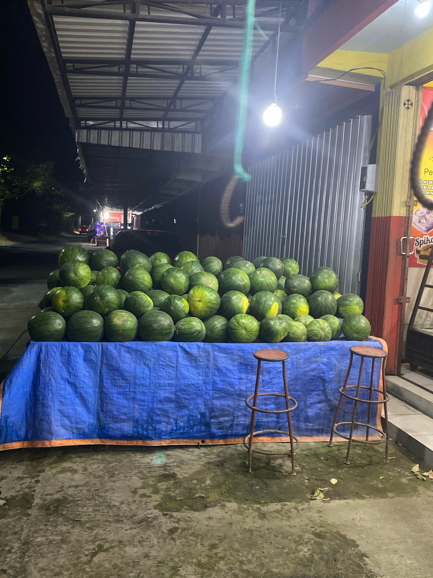 In Kediri City, Indonesia, at night, a vendor displays a large quantity of watermelons for sale. The watermelons are piled high on a table covered with a blue tarp, which is set up along the side of a building. Two rustic metal stools, likely for customers, are placed in front of the table. The area appears to be a roadside stall, with a corrugated metal wall and a covered awning suggesting it's a permanent or semi-permanent fixture. A bright light bulb illuminates the scene, casting a warm glow on the fruit and the surrounding structures. In the background, the street is visible, with faint lights suggesting other businesses or residences. The overall impression is a common sight of local commerce in a Southeast Asian city after dark.