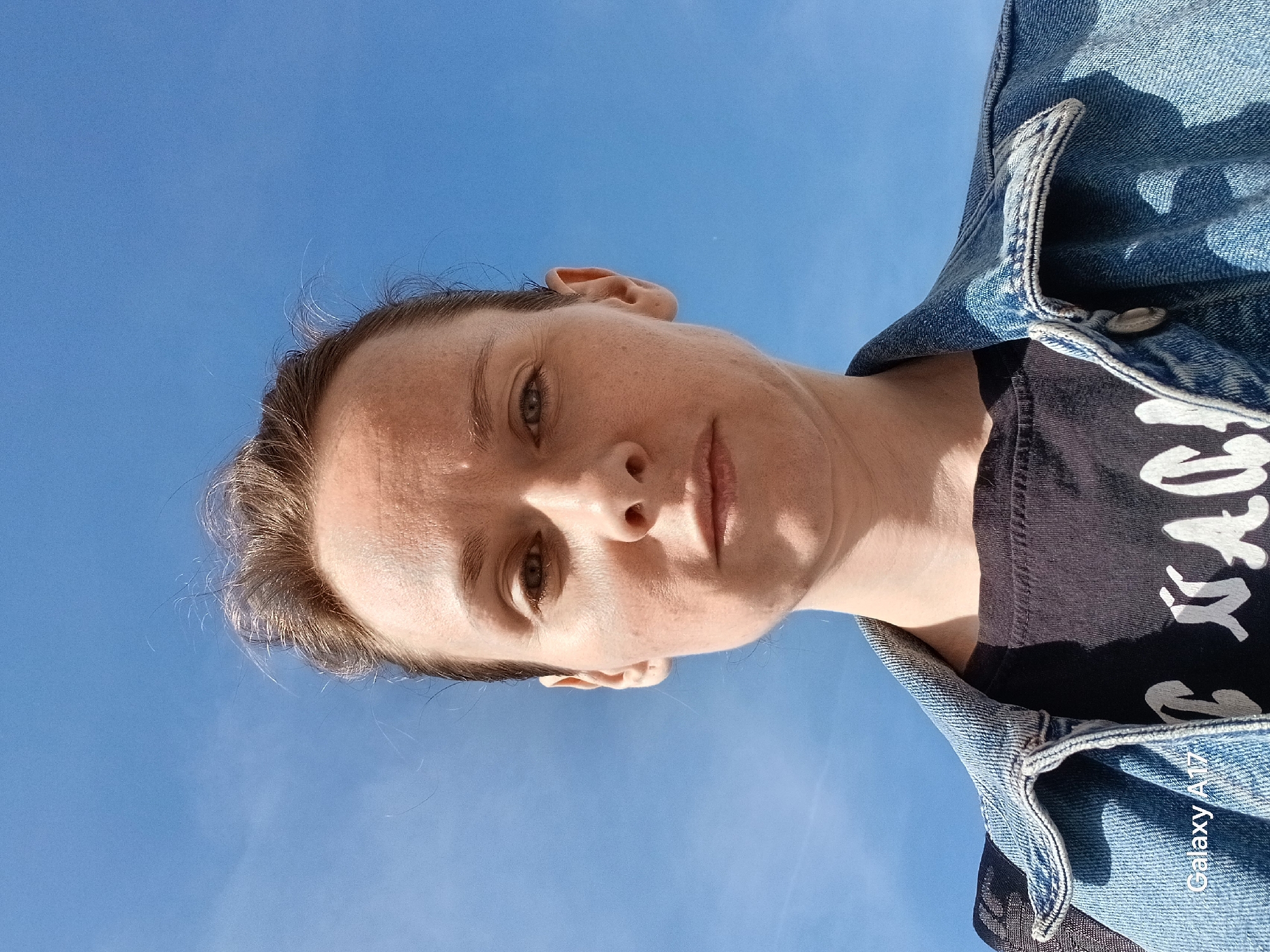 A low-angle portrait photograph captures a woman against a clear blue sky. The woman is wearing a dark-colored t-shirt and a denim jacket. The angle suggests the photograph was taken from below. There is no visible indication of the city Yekaterinburg, Russia. The photograph focuses solely on the individual and the sky.