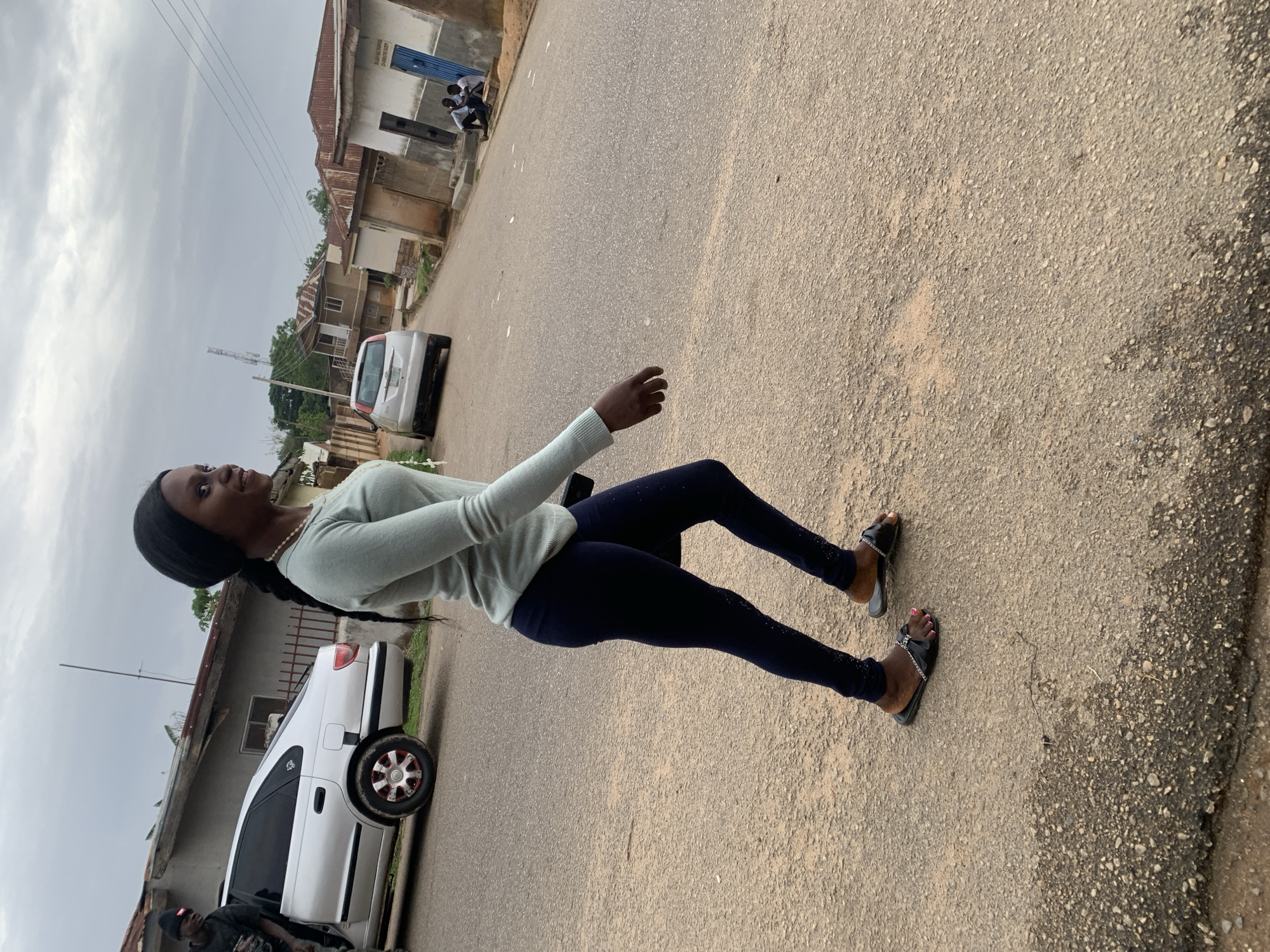 A young African woman stands on a paved road in Jos, Nigeria, wearing dark leggings, a light green sweater, and black sandals. She is looking up, with her arm outstretched. A white car is parked on the side of the road, and there are buildings in the background. The weather appears overcast. The scene is typical of a residential area in Jos.