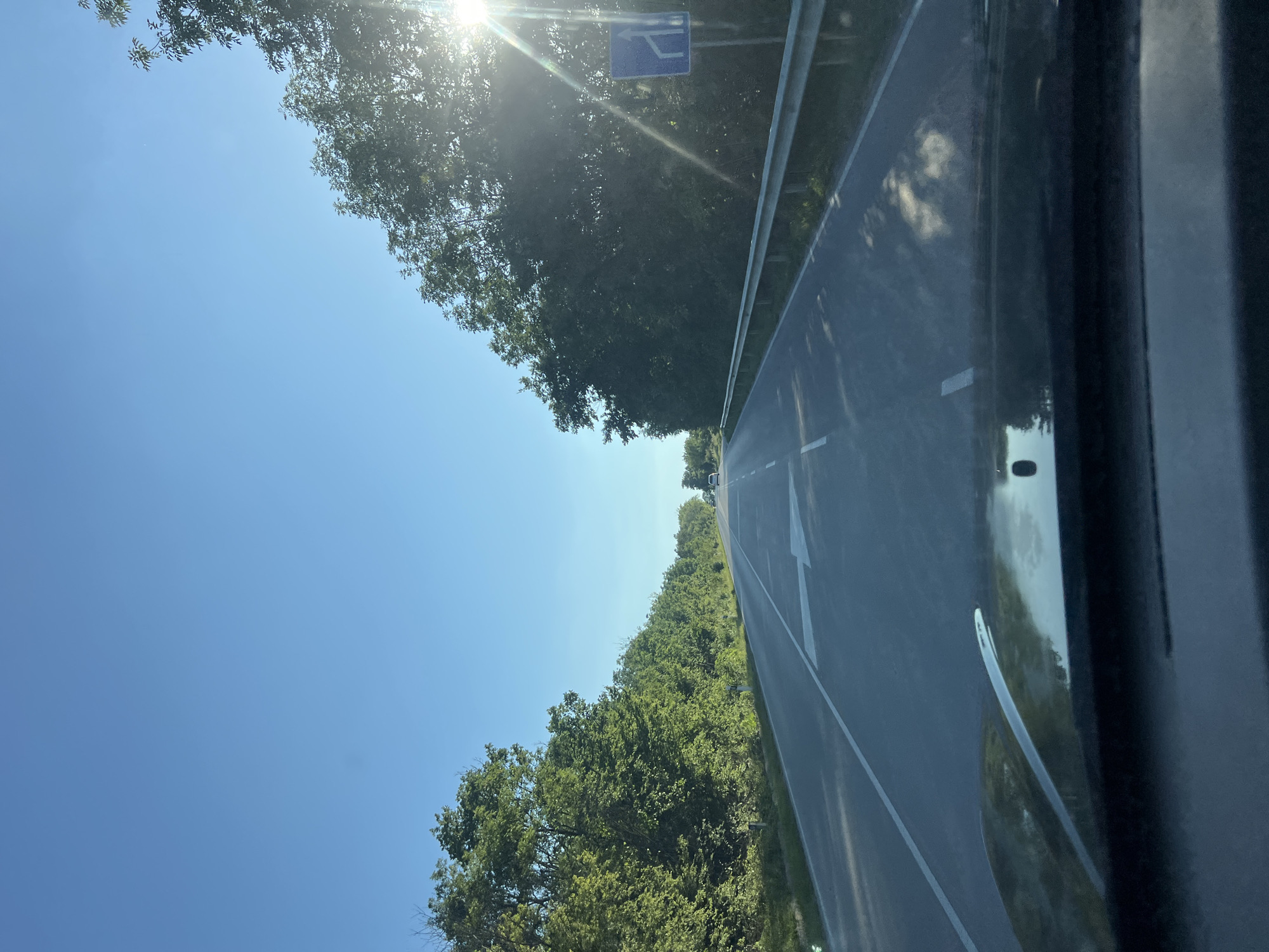 The image shows a view from inside a vehicle, looking out onto a road in Izvorche, Bulgaria. The road is paved and has white lane markings, including an arrow indicating the direction of travel. The roadside is lined with trees and vegetation. A blue traffic sign is visible in the distance near trees in the top center of the view. The sky is clear and blue, with the sun shining brightly. The vehicle's interior is partially visible in the foreground.