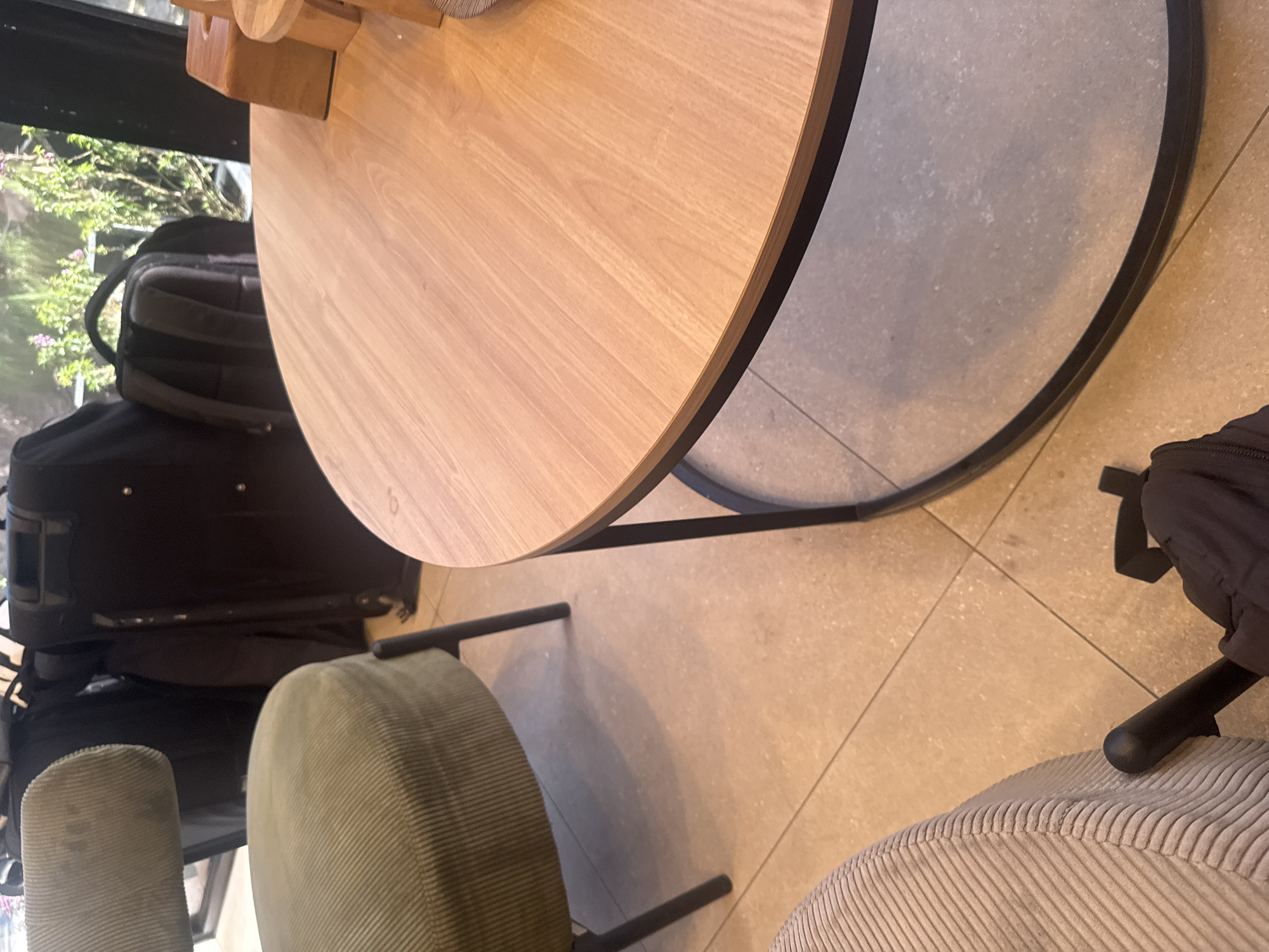 The image depicts an indoor scene, likely a cafe or lounge, with a light wood round table with a black metal base in the foreground. To the left, a portion of a green upholstered chair with a corduroy texture is visible. In the background, several dark suitcases and a black backpack are stacked, suggesting a travel hub or a place where people might be waiting for transportation. Beyond the luggage, a window offers a view of green foliage with some pink flowers, indicating an outdoor environment. The floor appears to be tiled in a neutral, sandy tone. The overall lighting suggests it is daytime. The scene is devoid of any people actively engaging in the space. There is no specific information in the image to suggest the location is Kuta Selatan, Indonesia.