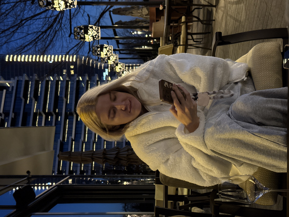 An image taken at dusk or night depicts a female with blonde hair seated outdoors on a patio. She wears a white plush zip-up jacket and grey casual trousers, looking down at a dark-colored smartphone held in both hands, interacting with its illuminated screen. A clear wine glass is visible on a dark table in the immediate foreground. The background features a row of distinctive decorative lanterns with patterned cutouts, providing warm illumination to an outdoor seating area with additional tables and chairs. Bare tree branches are silhouetted against a dark blue sky. Prominently in the background are multiple modern high-rise buildings, displaying illuminated windows and architectural lighting. One building exhibits horizontal lines of light, while a taller structure further back features a prominent vertical strip of illumination. The architectural style and urban setting strongly indicate the location as the Belgrade Waterfront development in Belgrade, Serbia.