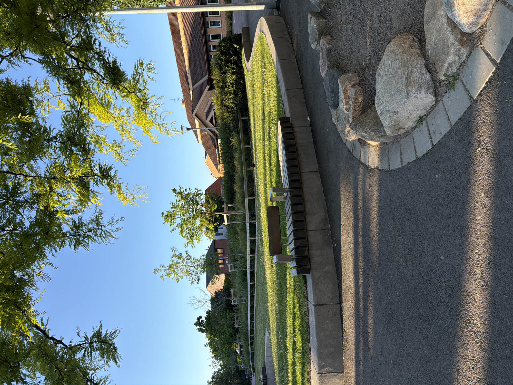 An eye-level, medium shot captures an outdoor seating area under a clear blue sky, framed by the lush, green foliage of trees. The scene is set on a sunny day, with sunlight casting long shadows across the asphalt ground in the foreground. In the mid-ground, a curved concrete planter bed encloses a patch of vibrant green grass. A modern bench, constructed from dark wood slats and metal supports, is positioned on the edge of the planter. Beyond the grass, a low wooden barrier runs along the perimeter of the area. In the background, several residential buildings with brown tiled roofs are visible, interspersed with trees and shrubs. A street lamp stands near the buildings. The overall impression is one of a well-maintained public space, likely a park or a community area, in a suburban or residential part of Helsingborg, Sweden.