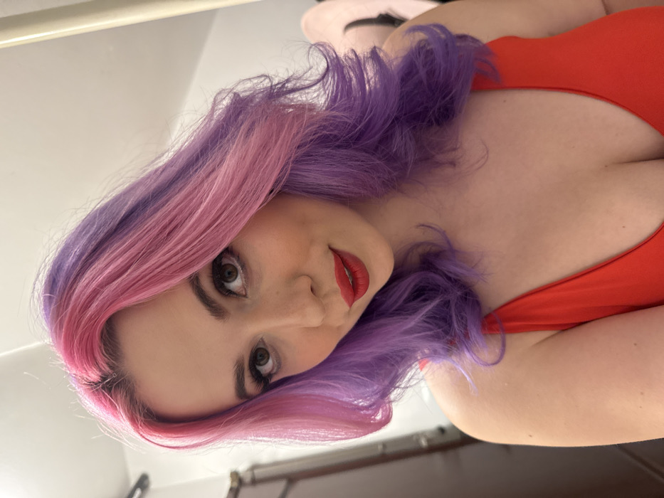 A woman with vibrant pink and purple hair reclines in a red garment. Her gaze is direct, lips painted a matching red. The angle suggests a low perspective, emphasizing her features and the texture of her hair. A glimpse of what seems to be a doorframe is visible in the background.