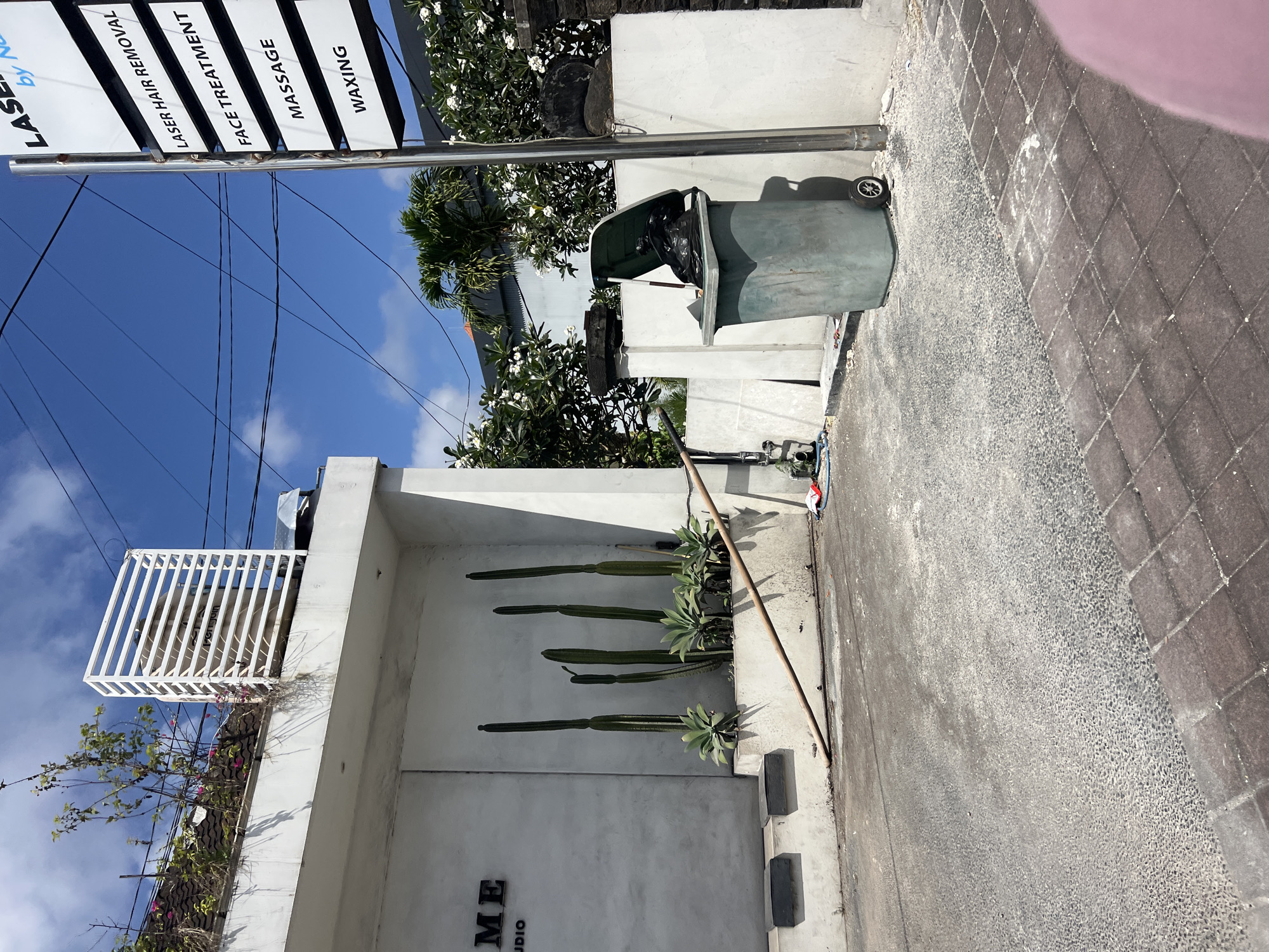 In Kuta Selatan, Indonesia, a green trash bin filled with black trash bags sits to the right of a white wall with a vertical sign that lists services: "LASER HAIR REMOVAL," "FACE TREATMENT," "MASSAGE," and "WAXING." The sign is mounted on a metal pole. To the left of the wall, a white building has the word "ME" in black lettering on its exterior, with "STUDIO" underneath. Several tall, thin cacti and smaller succulent plants are arranged in a planter box in front of the white building. A thin wooden pole lies on the ground, extending from the planter box towards the right. The ground in front of the building is paved with a rough, greyish material, and further right, a sidewalk is made of brown rectangular pavers. Overhead, a bright blue sky with scattered white clouds is visible, with power lines crisscrossing the frame.