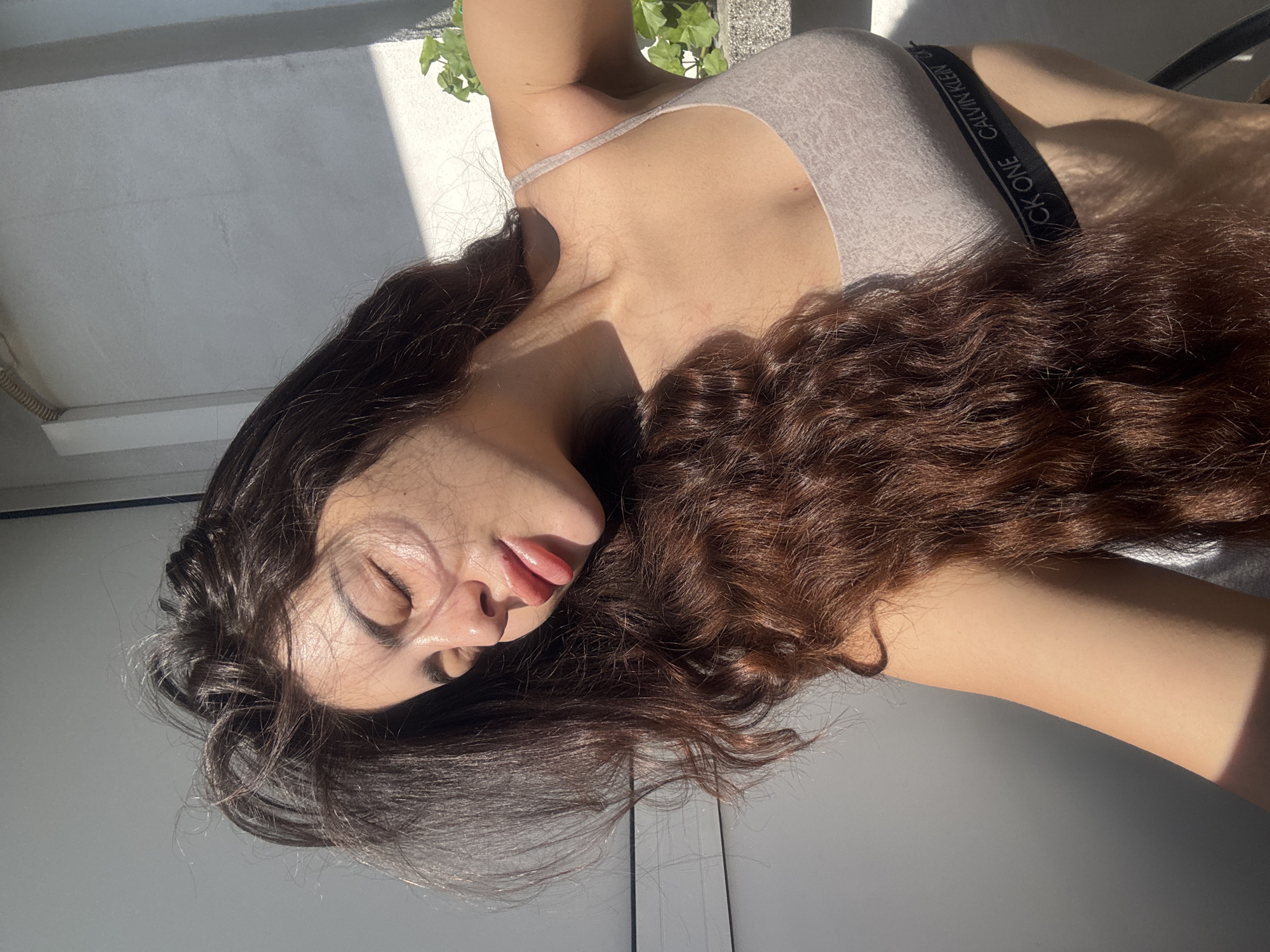 A medium shot, taken outdoors, portrays a woman with long, dark, wavy hair that transitions into a lighter auburn hue. Her eyes are closed, and she is lightly pursing her lips, showing a hint of her tongue. She is wearing a light grey Calvin Klein branded tank top. The scene is bathed in sunlight, creating stark shadows on her skin and the surrounding background, which includes a white wall, some greenery, and what appears to be a metal railing. The location is potentially a balcony or terrace in Plovdiv, Bulgaria.