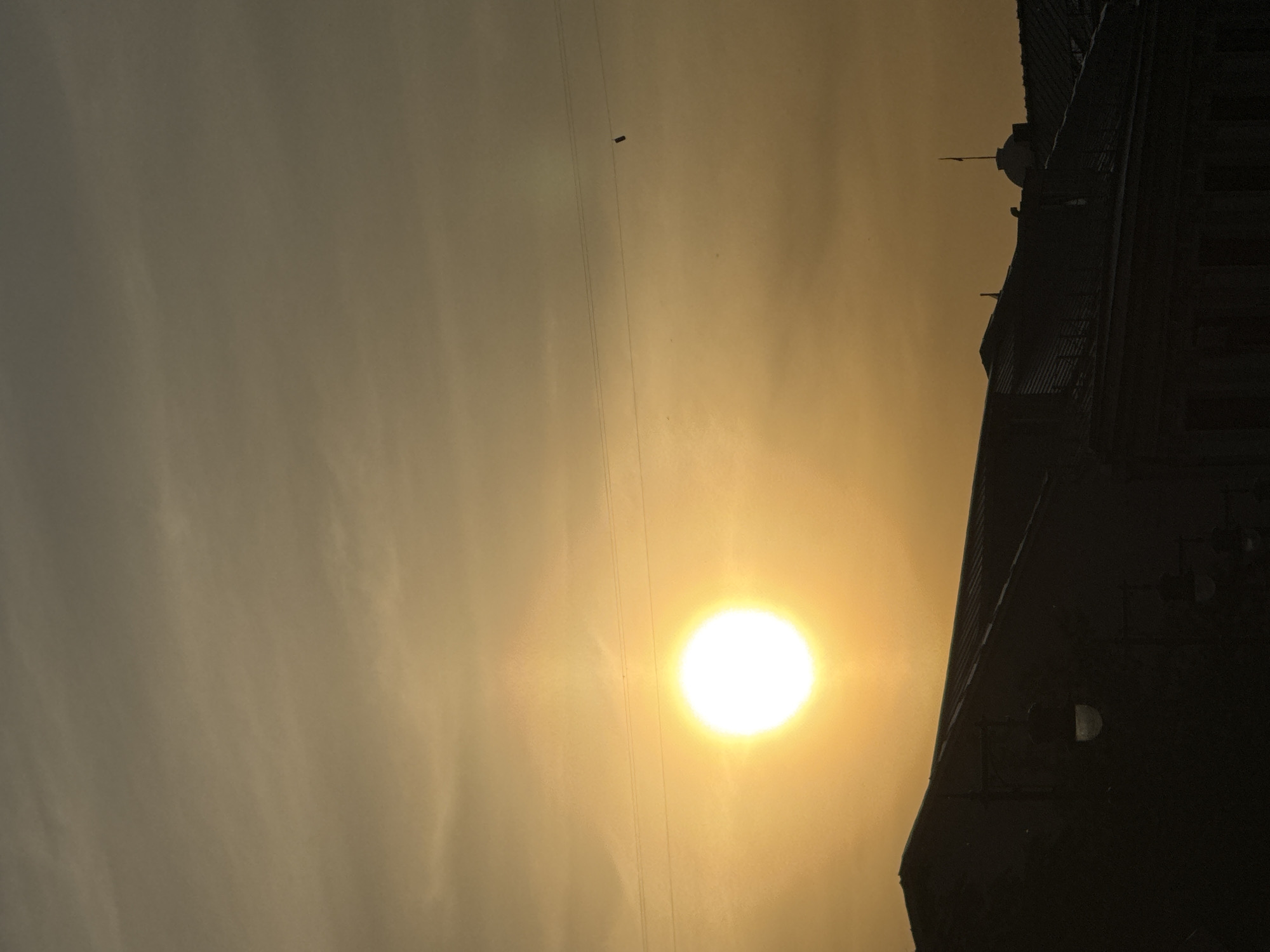 The image shows a hazy, golden sunset in Moscow, Russia. In the foreground, the silhouette of a building is visible on the right side. High tension electrical wires are visible running horizontally across the image. The sun is a bright white orb.