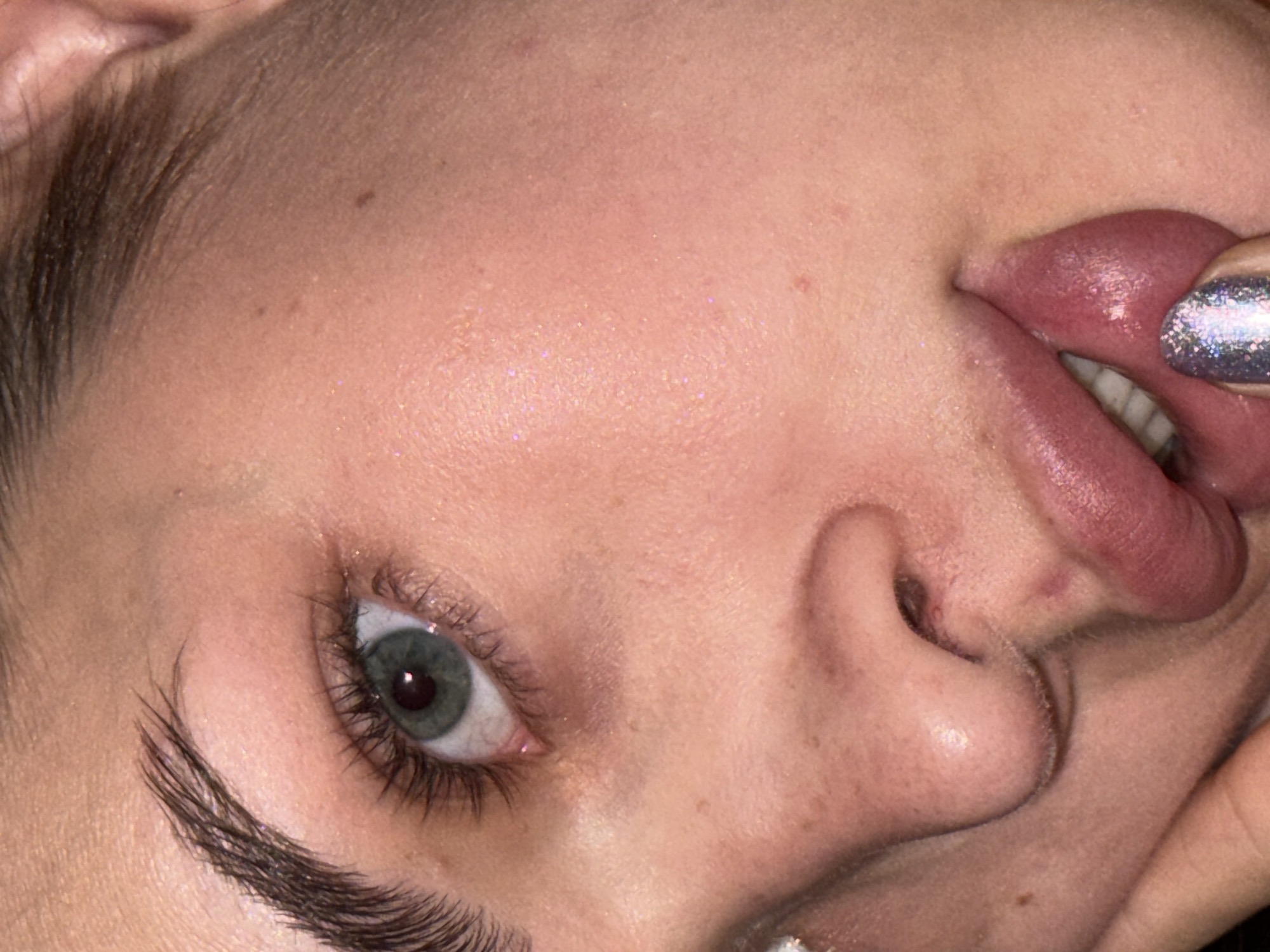 Close-up of a person's face, tilted to the side. The face includes an eye with long lashes, a nose, lips, and a portion of a finger with a glittery nail pressed against the lips. The skin has some shine. No location information is discernible from the image.