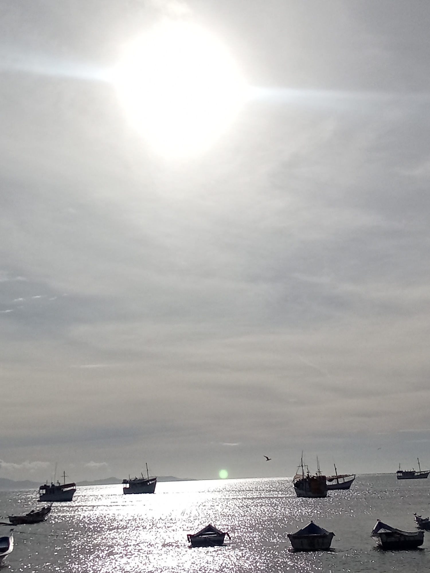 The image depicts a seascape in Juan Griego, Venezuela. The sun is high and obscured by clouds, creating a bright haze over the scene. The water is shimmering with sunlight. A number of boats, some small, some larger, are scattered across the water. A bird flies in the distance. The coastline is visible in the background.