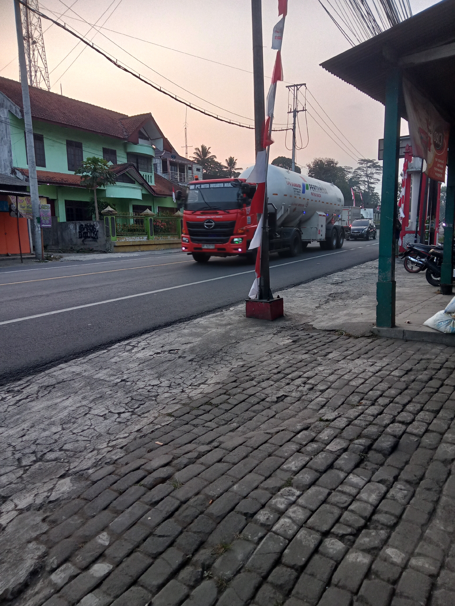 An exterior, medium-angle, daylight shot captures a street scene in Tegalrejo, Indonesia. In the foreground, a rough, cracked asphalt and cobblestone pavement leads to a road. A red truck with a white tanker trailer, emblazoned with the "PERTAMINA" logo, is driving down the road. The truck has Indonesian flags attached to a pole in front of it. On the right side of the road is a covered walkway with green posts. A motorcycle is parked under the walkway. On the left side of the road are green buildings with red-tiled roofs, a tall tower, and trees. The sky is a pale orange, suggesting it is either sunrise or sunset.