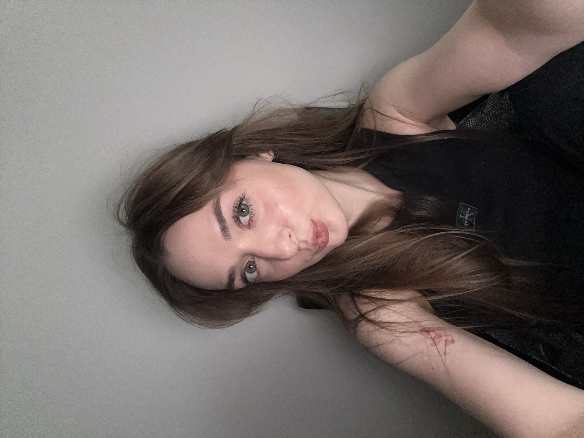 A woman with long brown hair is posing for a selfie. She is wearing a black tank top with a small square logo on the upper left chest. The background is a plain gray wall. There appears to be a small reddish mark or scratch on her left forearm. The location of Kragujevac, Serbia is not evident in the image.