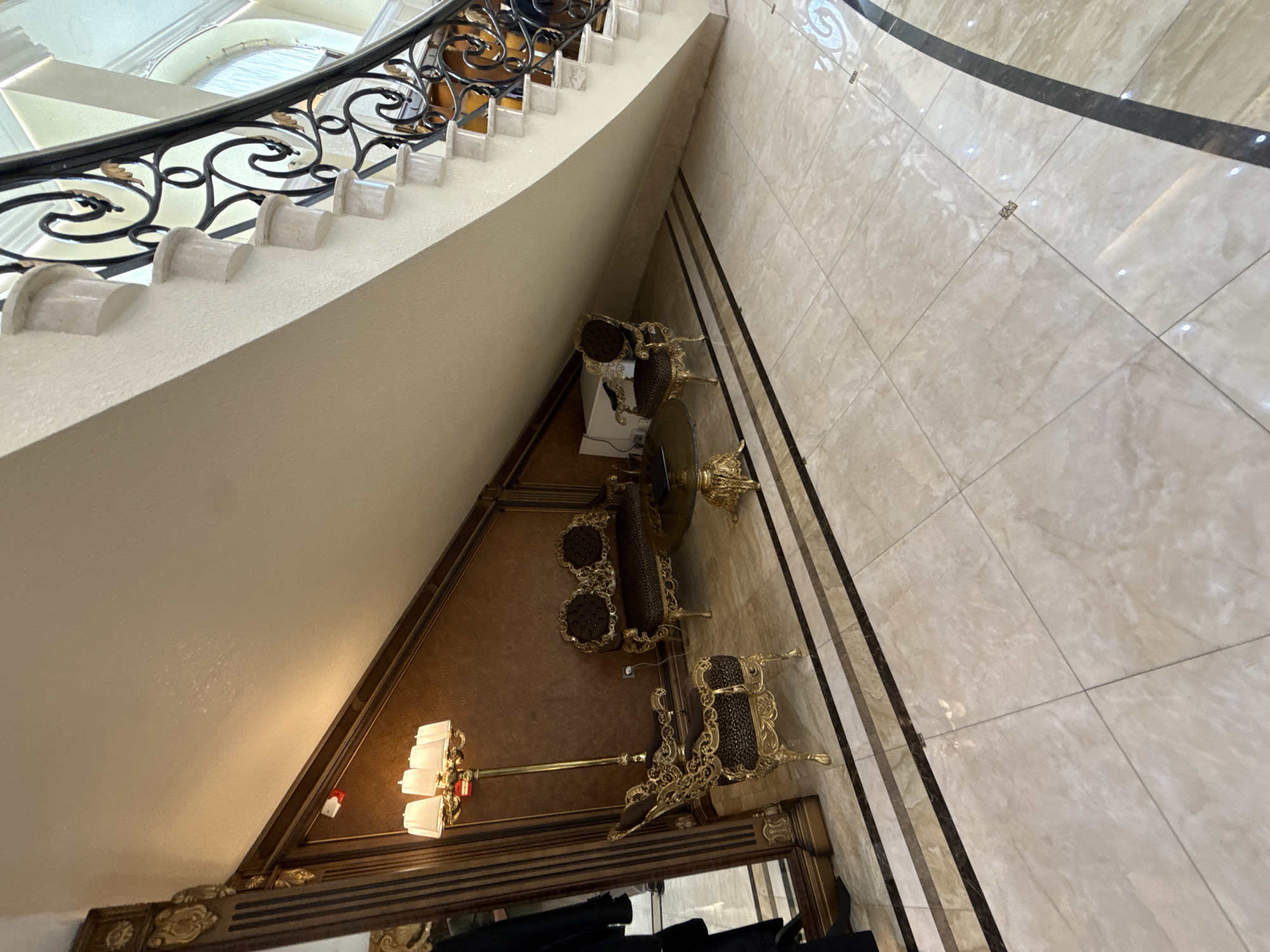 The image depicts a luxurious interior space, likely a foyer or grand hall, with a prominent staircase. The flooring is a large-format, polished beige marble with inlaid decorative borders. To the left, under the staircase, is a seating area featuring a gold-accented sofa with dark upholstery and a matching armchair. A floor lamp with multiple shades stands nearby. A tall, ornate mirror is visible to the far left, reflecting a portion of the room and a person standing by. The staircase itself is grand, with white marble steps and a black metal balustrade featuring ornate gold scrollwork. The walls in this section are paneled in a rich, dark wood tone. In the distance, the hallway curves, with more marble flooring visible. The overall impression is one of opulence and classic design. This setting is consistent with the high-end residential and commercial architecture found in Baku, Azerbaijan.