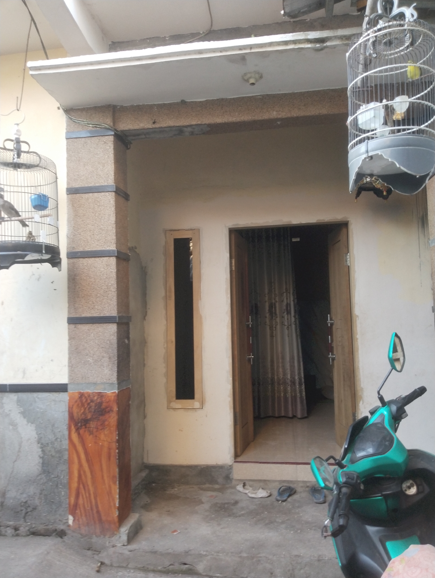 A motorcycle, possibly a Honda Beat, is parked on a concrete surface in front of a house. The motorcycle is green and black and has a side mirror visible. A birdcage with two birds, one yellow and one brown, hangs from the porch ceiling. Another birdcage, with at least one bird, is also visible. The house has a wooden door and a narrow window. A tall, square pillar with a wood-grain pattern supports the porch roof. Several pairs of sandals are scattered on the ground. The image appears to be taken during the day.