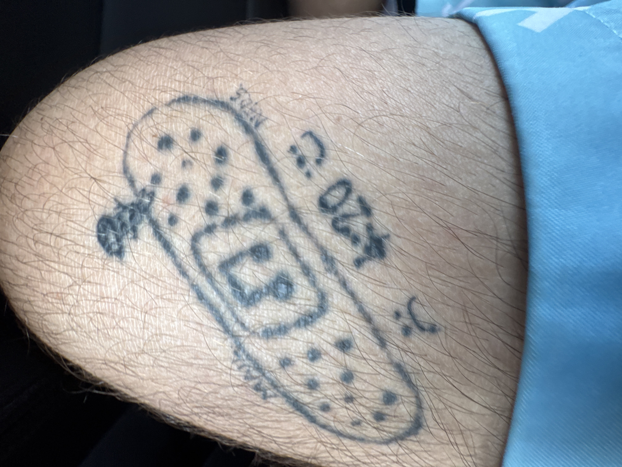 The image shows a close-up of a tattoo on a person's leg. The tattoo depicts a band-aid with dots, a musical note, and the text "4:20". There are also small smiley and frowny faces drawn near the text. The person is wearing a blue shirt. The image is taken from a high angle, looking down at the leg. There is no information or indication of the city of Chiari, Italy in the image.