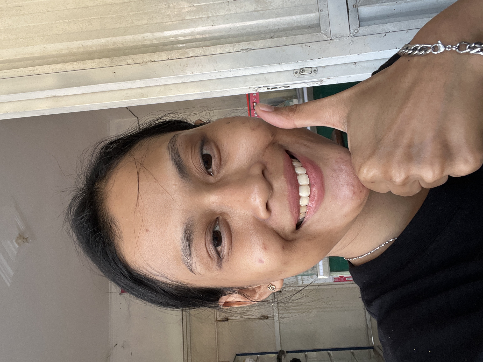 A young Indonesian woman of East Asian descent, with dark hair pulled back and fair skin, smiles and gives a thumbs-up gesture while standing indoors. She is wearing a black t-shirt, a silver necklace, and a silver bracelet on her right wrist. Her left earlobe has a small, gold earring in the shape of a flower. The background is out of focus, but appears to be a room with white walls and a metal door on the right. The lighting suggests it is daytime. The photo was taken in Kecamatan Pati, Indonesia.