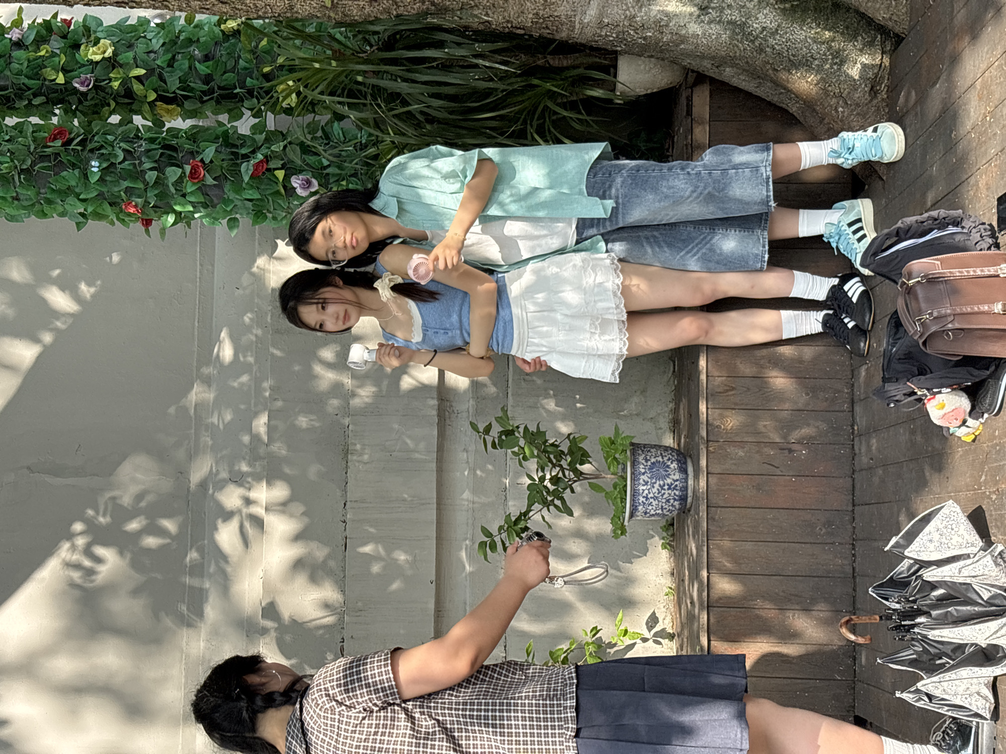 This image depicts a candid outdoor scene in the Yuzhong District of China. Three individuals are present. On the left, a person with dark braided hair, wearing a brown and white checkered shirt and a dark blue pleated skirt, is holding a camera and appears to be taking a photo or video of the other two individuals. The other two individuals are standing closer together, facing the camera. The person on the left of this pair has long dark hair, wears a blue sleeveless top with white lace trim, and a white ruffled skirt. They are holding a small white fan. The person on the right has dark hair, wears glasses, and is dressed in a light blue checkered shirt over a white top, with loose-fitting blue jeans. This person is holding a pink fan. They are standing in front of a textured concrete wall with dappled sunlight and shadows, and to the right, a large tree with lush green foliage and faux flowers. In the foreground, on a wooden deck, there is an open black and white umbrella and several bags, including a brown leather satchel. A potted plant with blue and white oriental-style ceramic is visible on a wooden ledge behind the individuals.