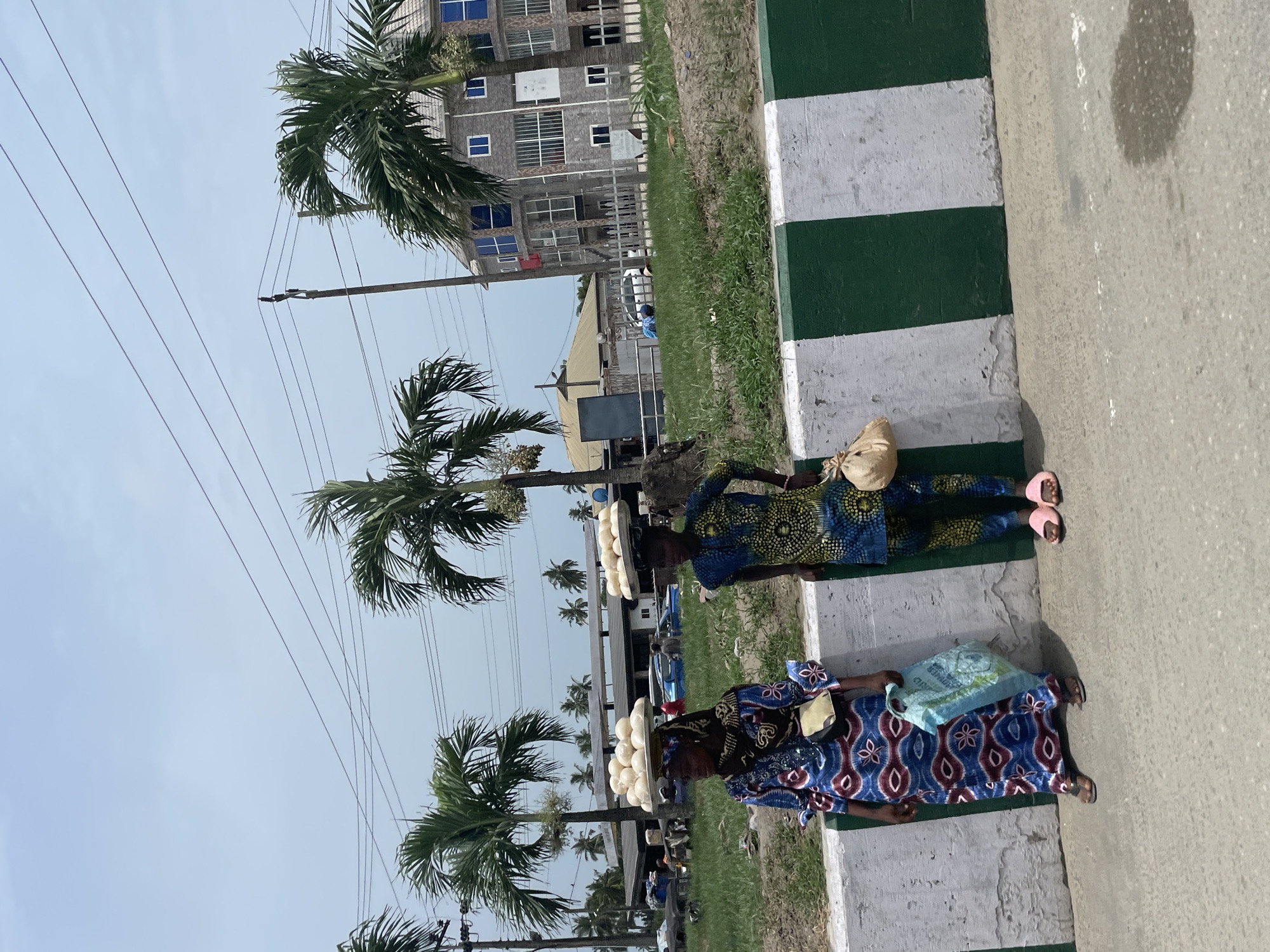 Two women stand on a sidewalk in Badagry, Nigeria, each balancing a tray of white, round food items (possibly bread or snacks) on their heads. The woman closer to the camera wears a blue and red patterned dress and holds a light blue shopping bag. The other woman wears a blue and yellow patterned dress and carries a tan-colored bag. Both wear sandals.
They are standing adjacent to a curb painted with alternating white and green stripes. Palm trees line the street, and a multi-story building is visible in the background. Overhead power lines run across the sky. The scene appears to be a typical day on a Badagry street with women selling goods.