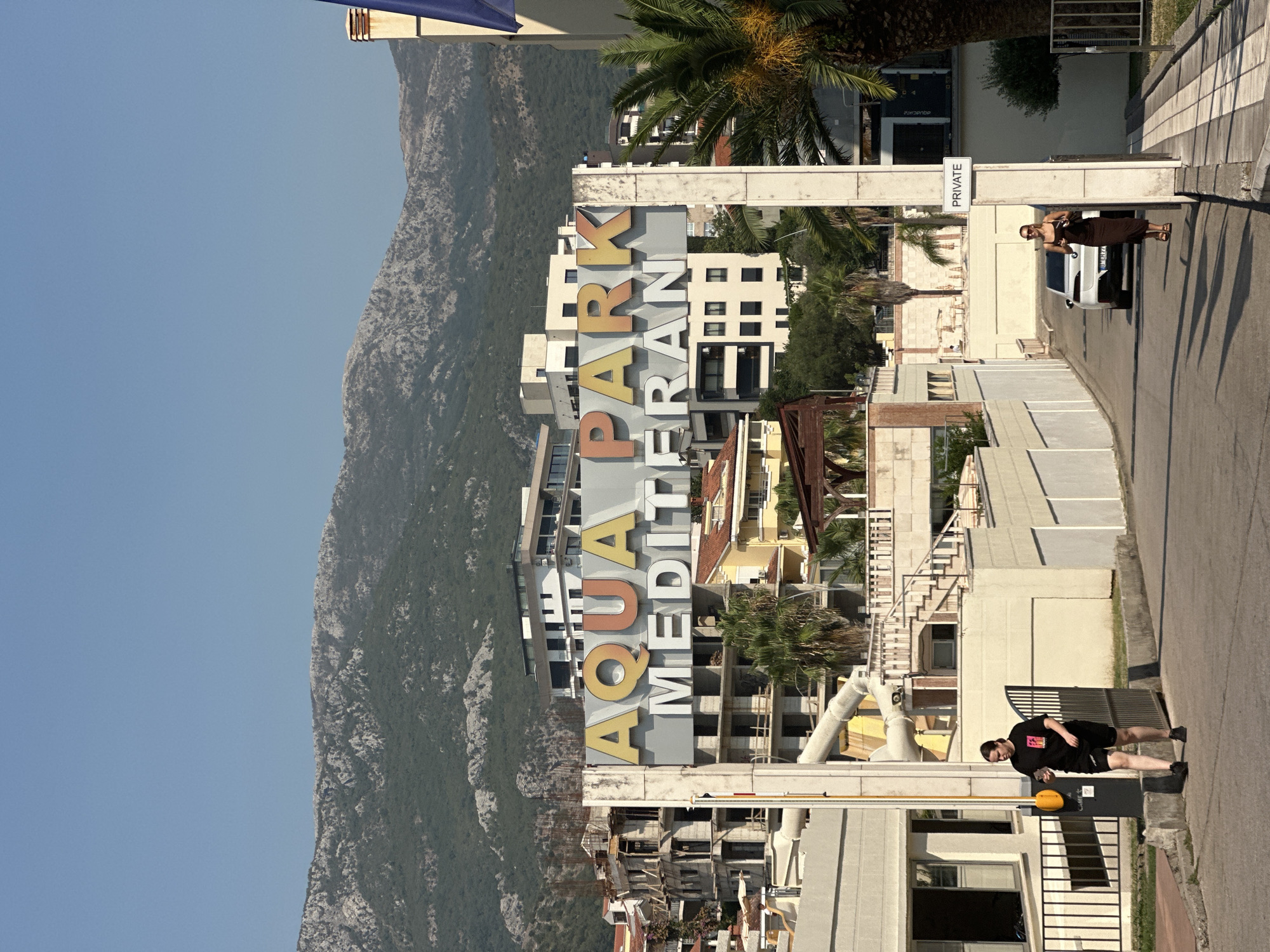 An eye-level, medium shot captures the entrance to Aqua Park Mediteran in Bečići, Montenegro. The sign for the aqua park, with colorful "AQUA PARK" lettering in yellow and orange and "MEDITERAN" in white, is prominent. Behind the sign, a partially constructed building and other modern buildings rise against a backdrop of green, mountainous terrain under a clear blue sky. In the foreground, a person in a black t-shirt and shorts walks away from the camera on the left side of the asphalt road. A white car drives past a person in a brown dress walking towards the camera on the right. A "PRIVATE" sign is mounted on a pole near the right edge of the frame. Palm trees and other vegetation are visible throughout the scene.