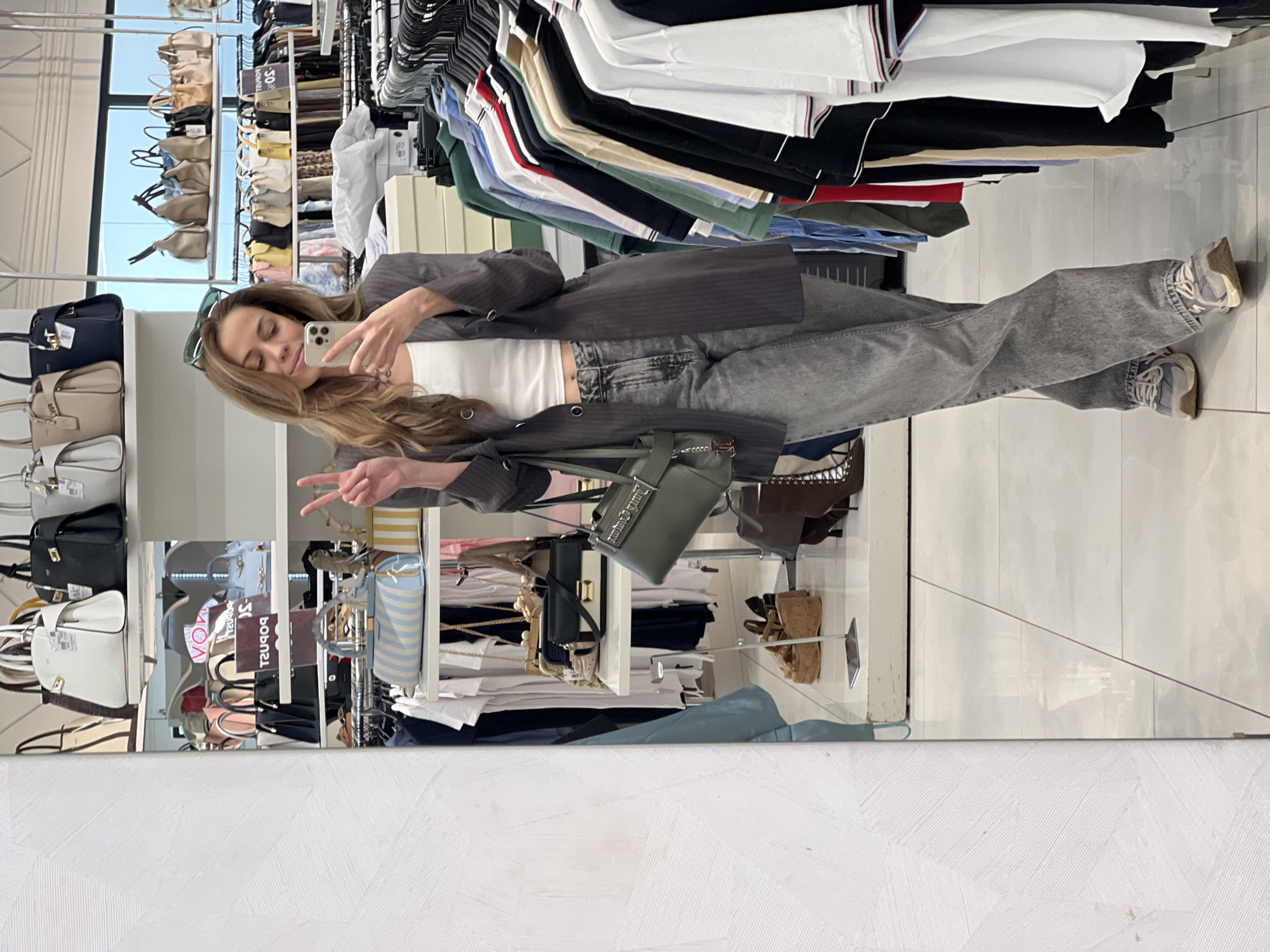 A young woman stands sideways in front of a large mirror in a clothing store, possibly in Budva, Montenegro. She is taking a photo of herself with her smartphone, making a peace sign with her left hand. She wears a white crop top, a gray pinstripe blazer, wide-leg gray jeans, and gray sneakers. A dark green handbag hangs from her shoulder. Sunglasses are perched on her head. The background shows racks of clothes and bags. The floor is light-colored tile. The bottom of the image reveals the mirror's edge against a white textured wall.