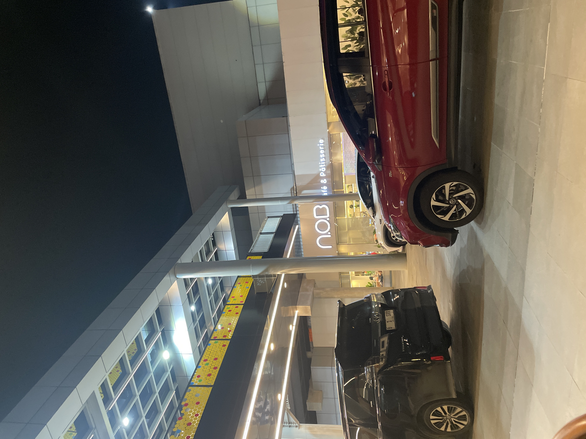 The image shows an outdoor scene at night in North Jakarta, Indonesia, near a building with a storefront labeled "n.o.b. Café & Pâtisserie". Two parked cars are visible: a red SUV and a black pickup truck. The building features a modern design with multiple levels and illuminated windows, suggesting a commercial or shopping complex. The setting is well-lit, with artificial lights highlighting the architecture and parking area.