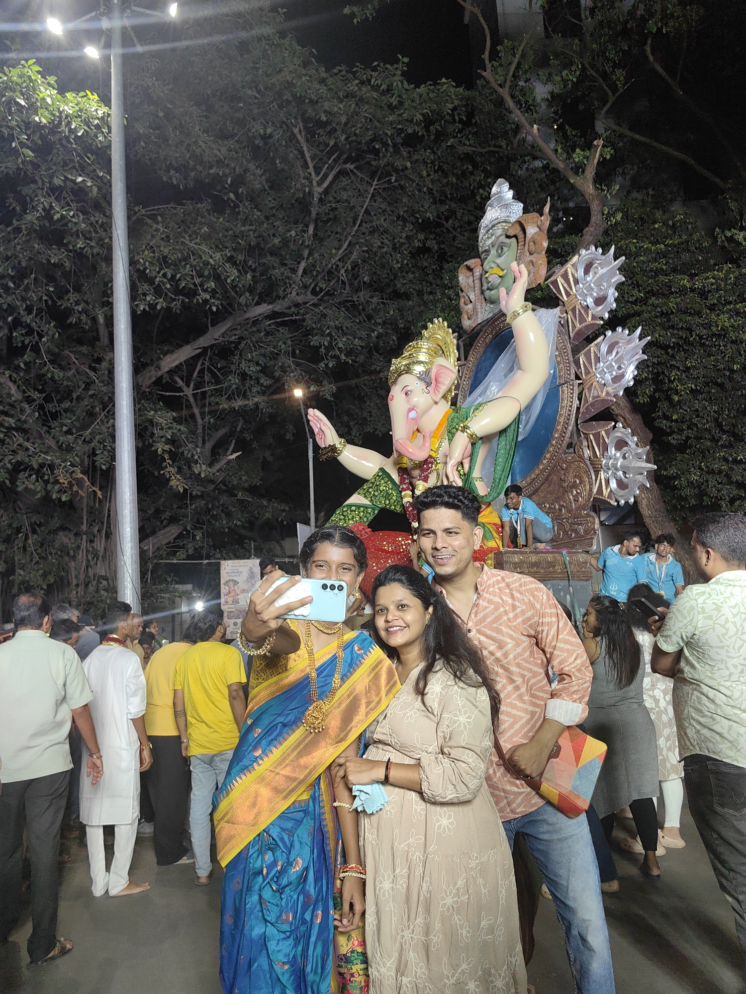 The image captures a street scene in Mumbai, India, at night, likely during a religious festival given the presence of a large Ganesha idol. In the foreground, three people are taking a selfie. A young woman, dressed in a blue and gold saree with elaborate jewelry, holds up a light blue smartphone. Beside her, a man in a patterned orange and white shirt and blue jeans smiles at the camera. To their right, another woman in a beige, floral-patterned dress also poses for the photo, holding a light blue mask. In the background, a colossal statue of Lord Ganesha, adorned with vibrant colors and intricate details, towers over the scene. Several other people, some in traditional Indian attire and others in casual wear, are visible, suggesting a public gathering or procession. The environment is dimly lit by streetlights, with lush green trees surrounding the area.