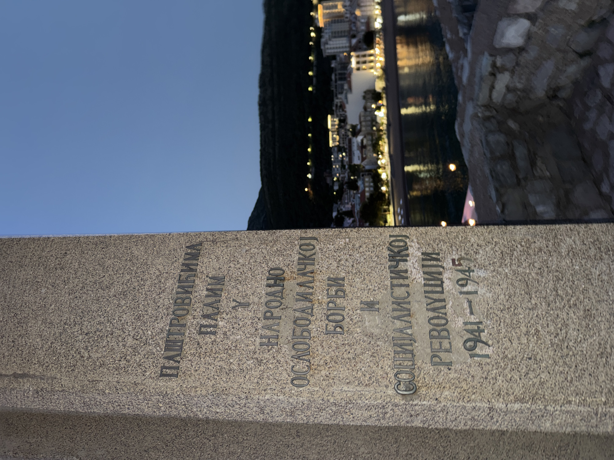 The image depicts a commemorative stone monument in Gornji Katun, Montenegro. The inscription on the monument is in Cyrillic script and reads: "PAŠTROVIĆIMA PALIM U NARODNO OSLOBODILAČKOJ BORBI I SOCIJALISTIČKOJ REVOLUCIJI 1941-1945". In the background, one can see a coastal city illuminated at night, reflecting in the water. To the right of the monument is what seems to be part of a stone structure.