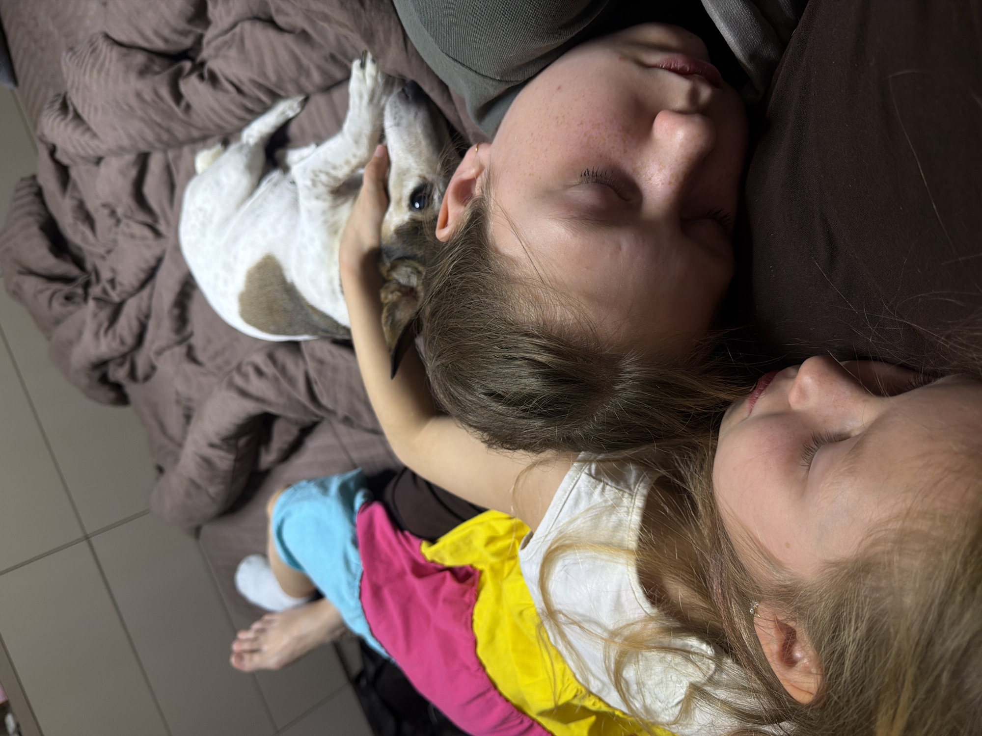 A slightly elevated, close-up shot shows a young adult lying on a bed with a child and a dog. The young adult is on their side, facing left, with their eyes closed. They are wearing a dark-colored top. A child is lying next to them, also with closed eyes, facing right. The child is wearing a white shirt with layers of yellow, pink, and blue clothing underneath. A dog is lying on the bed near the adult's head, on a brown bedspread. The dog is white with brown markings. The background includes a tile floor in shades of tan and brown. It is not possible to verify the location in Dubovaya, Russia from the image.