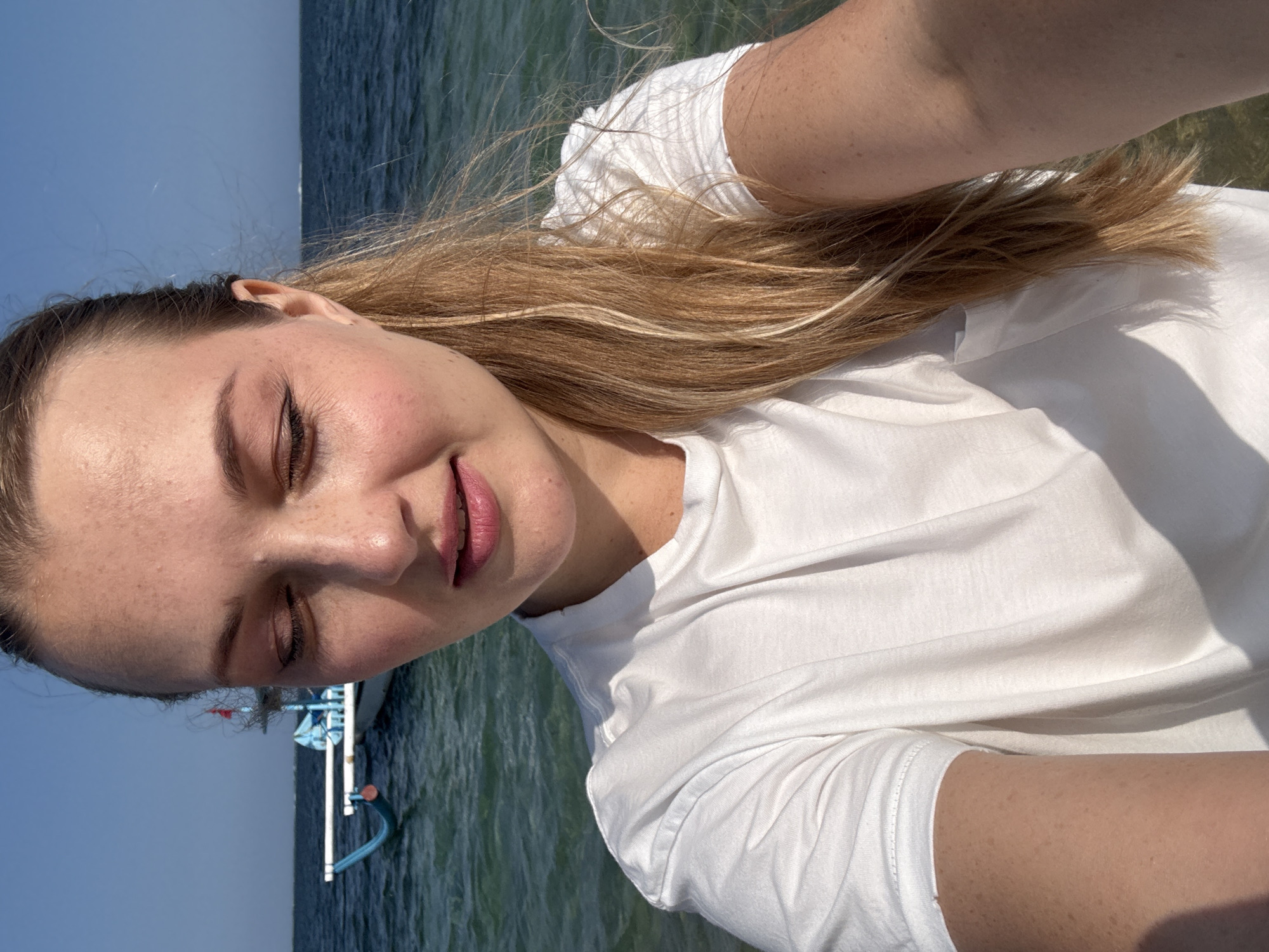 A woman with blonde hair pulled into a ponytail stands on a beach, looking down. She wears a white t-shirt. Behind her, the ocean is visible, with a blue and white boat in the distance. The sky is clear and blue. The image appears to have been taken in Denpasar, Indonesia, given the presence of a traditional Indonesian fishing boat.