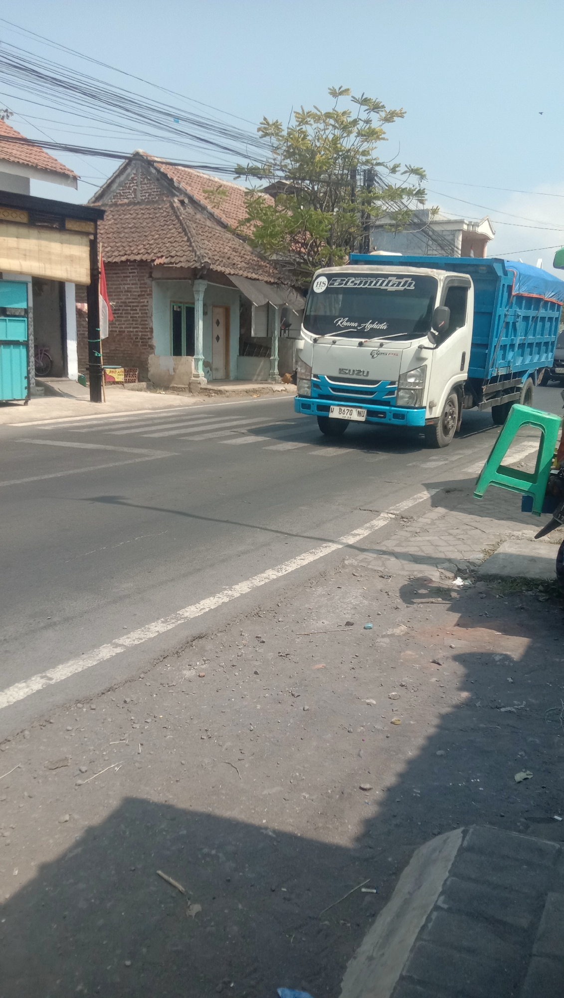 The image shows a street scene in Karanganyar, Indonesia, under a clear blue sky. A white and blue Isuzu truck is parked on the right side of the road, near a pedestrian crossing. The truck has a blue tarp covering its cargo bed. In the background, there are traditional Indonesian houses with tiled roofs and a tree with green foliage. There are also power lines crisscrossing the sky. On the left side of the road, there is a building with a turquoise facade. The road surface is asphalt with white markings for the pedestrian crossing. A green plastic chair is placed on the sidewalk to the right of the truck. The overall impression is that of a typical street scene in a semi-urban area in Indonesia.