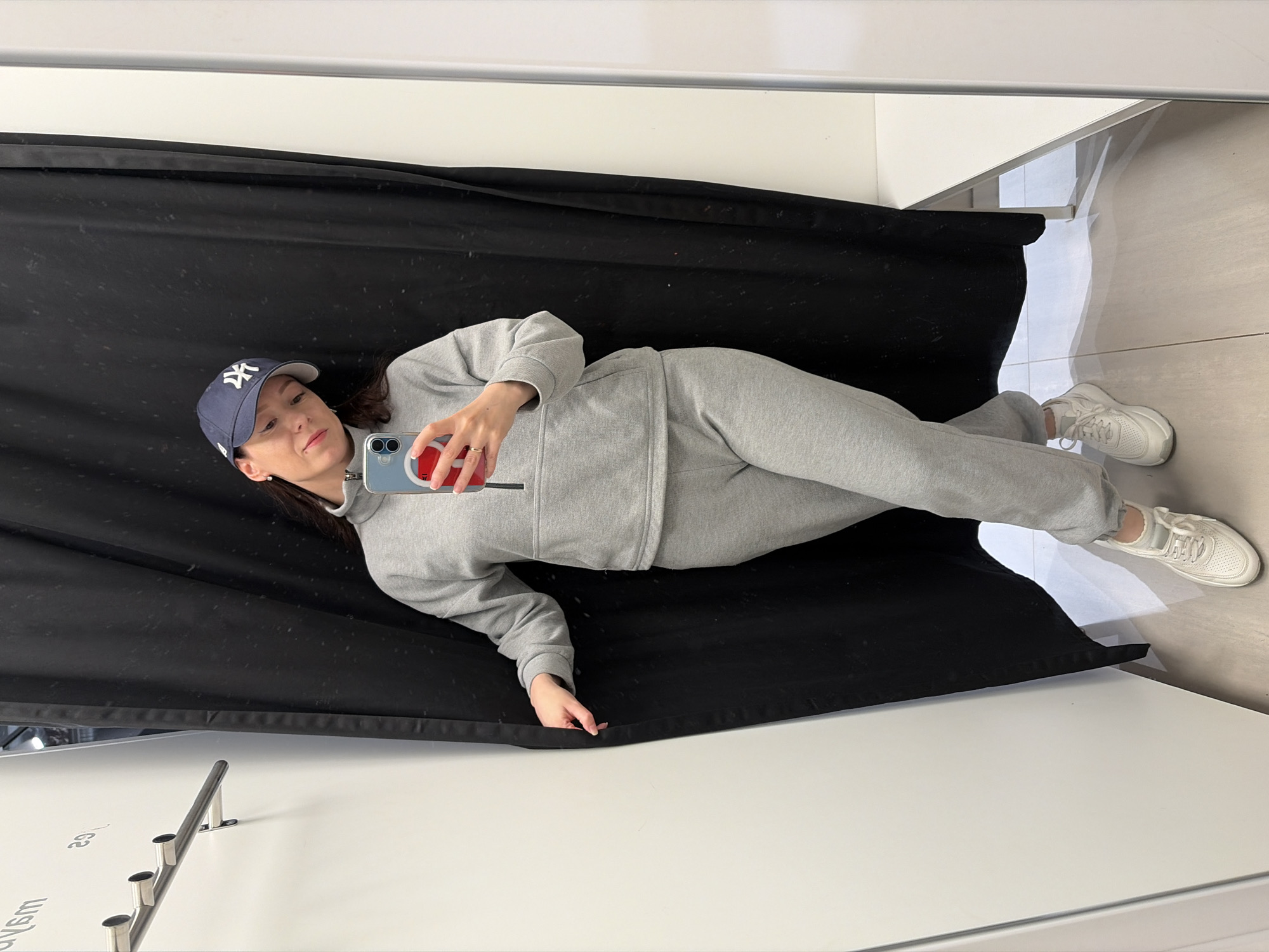 A young woman is captured in a mirror selfie, positioned horizontally across the frame. She wears a casual grey sweatsuit, comprised of a hooded sweatshirt and matching sweatpants, paired with white sneakers. A dark blue baseball cap with a white "NY" logo is worn on her head. She holds a light blue/grey smartphone, adorned with an orange pop socket or grip, in her right hand, directing her gaze towards its screen. A ring is visible on her left ring finger, and she wears small earrings.
The setting appears to be an indoor space, possibly a dressing room, studio, or retail area. A large black fabric curtain or backdrop runs behind the woman, providing a strong contrast to the white tiled floor and pristine white walls or surfaces. A metal rod with three cylindrical attachments, potentially a clothing display, is visible on one of the white surfaces in the foreground. The image's orientation implies it's a reflection, with the woman lying parallel to the ground. Inverted text, including "mб\40" and "гз," is faintly visible on a white surface, suggesting labels in a commercial setting. The indoor environment provides no specific cues for time of day or weather. This scene is set in Япрыково, Russia.