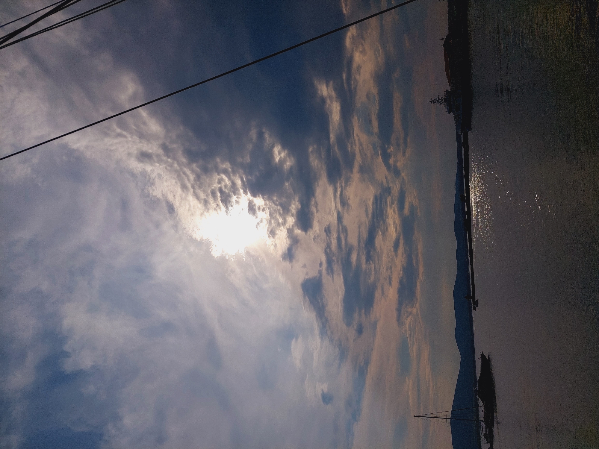 The image depicts a body of water with a silhouetted cityscape in the background, under a dramatic cloudy sky. The sun is visible through the clouds, casting a bright glow and creating highlights on the water's surface. On the left, a sailboat is anchored. Further in the distance, a larger vessel, possibly a ship, is docked near a building on the right side of the frame. The scene appears to be a harbor or marina, likely in Sozopol, Bulgaria, given the presence of traditional architecture visible in the distant cityscape and the common boat activity in the area. The overall atmosphere is serene, with the natural elements of the sky and water dominating the view.