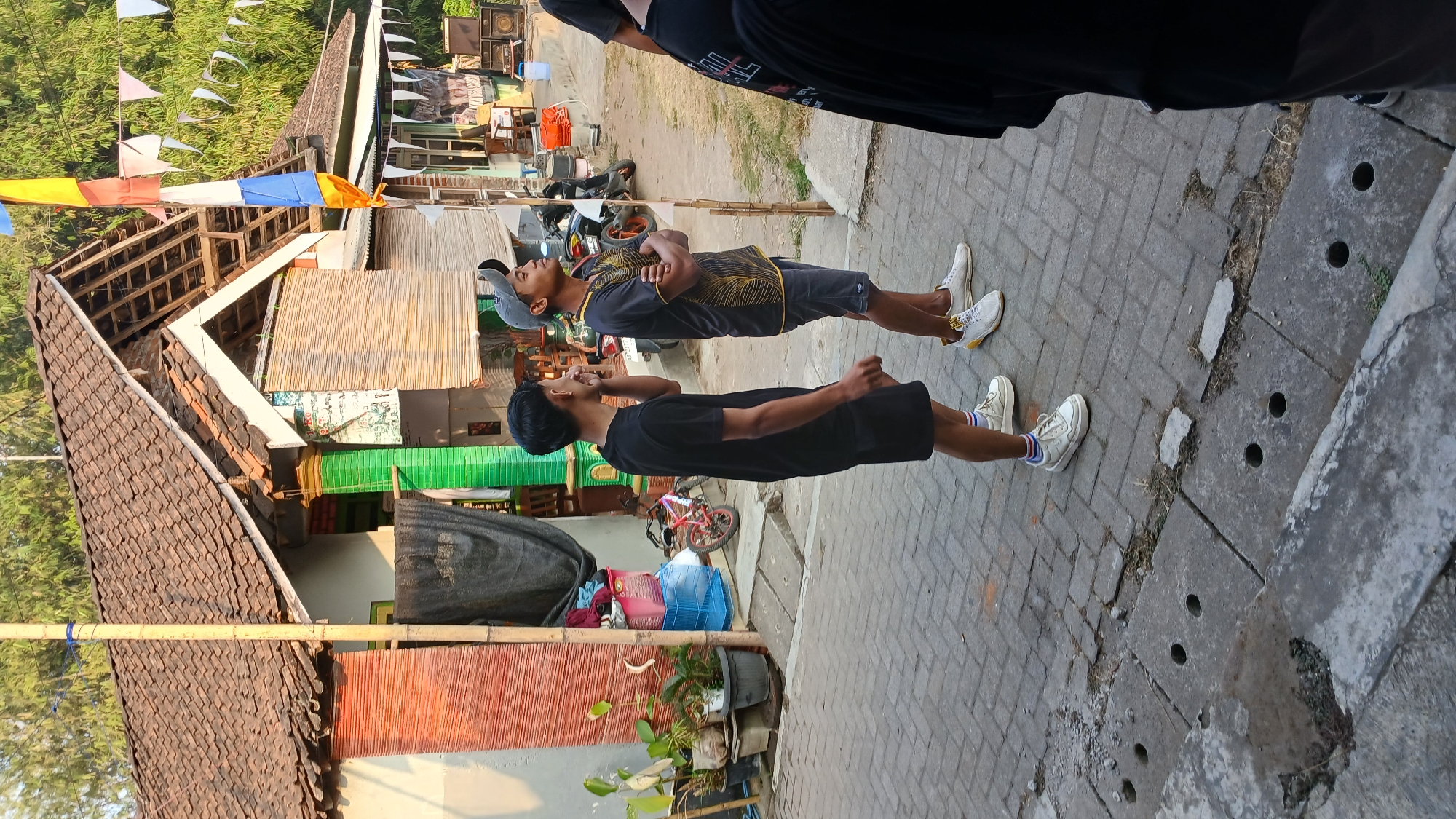 In Mendut, Indonesia, two young men stand on a paved street in front of a traditional house with a tiled roof. The man on the left wears a black t-shirt and shorts, while the man on the right is dressed in a black t-shirt with yellow accents, grey shorts, and a grey baseball cap. They appear to be looking upwards and conversing. Behind them, the house features bamboo blinds on its windows and colorful decorations, including flags. Various objects like potted plants, a bicycle, and audio equipment are scattered around the scene. The environment suggests a casual outdoor gathering or event taking place.