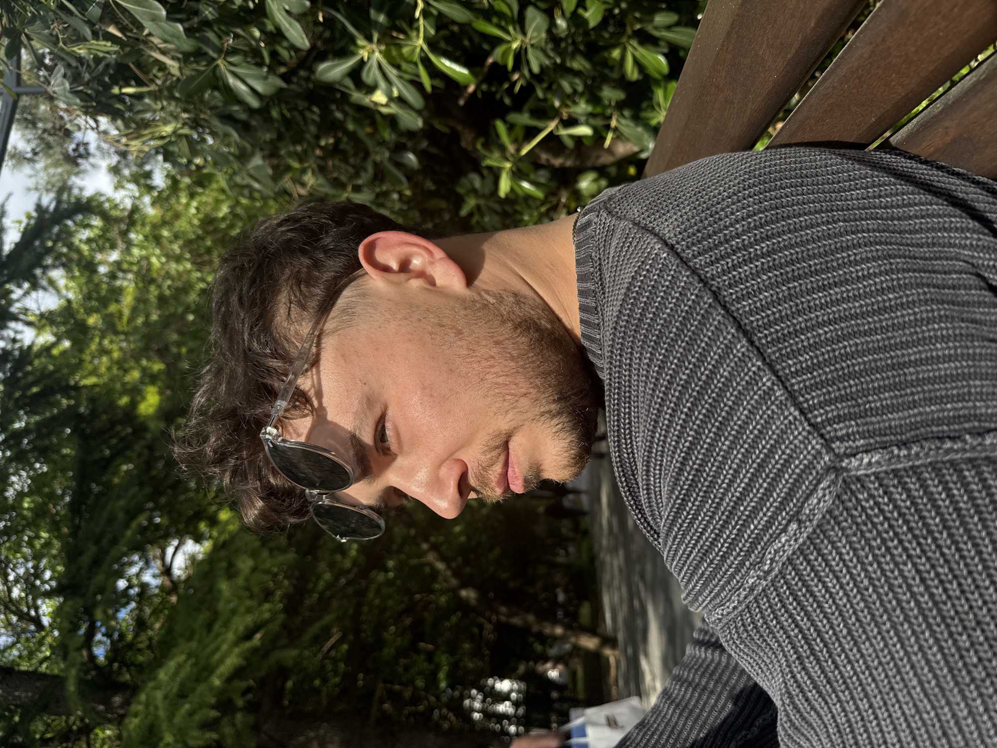 A man with short dark hair and a beard sits on a wooden bench. He wears a gray sweater and sunglasses that are partially tucked into his hair. He is looking down and to his left. The background consists of green foliage. The location is likely in Bar, Montenegro, given the prompt instructions.