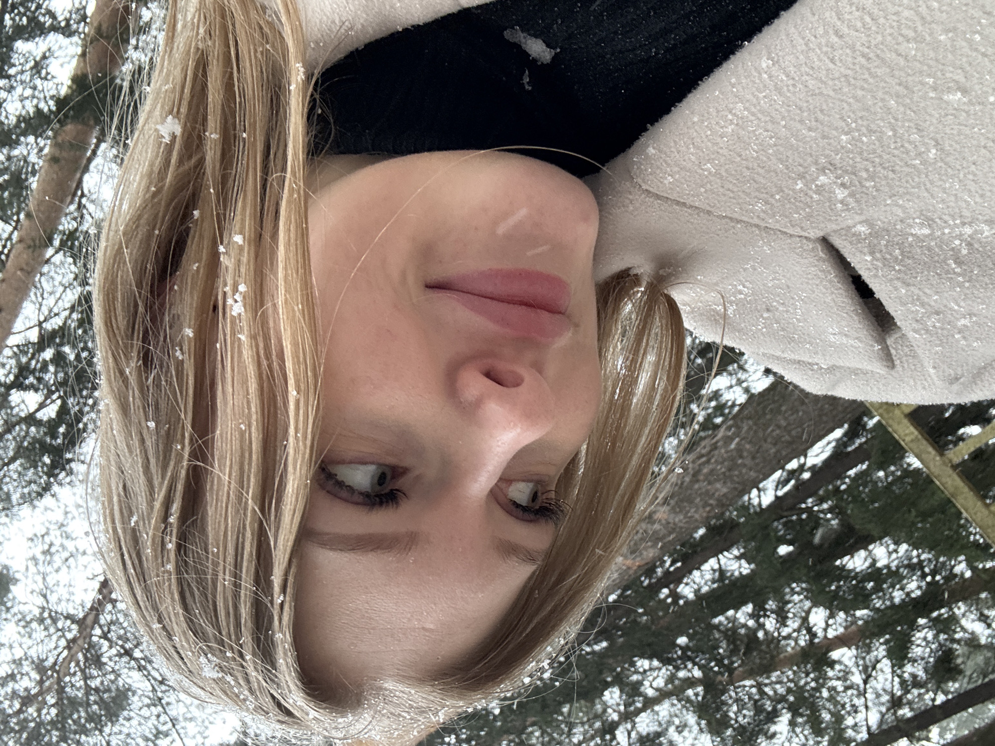 The image is a close-up, upside-down shot of a blonde woman with snow in her hair. She is wearing a black turtleneck and a light-colored coat covered in snow. The background shows trees covered in snow, suggesting a snowy environment. Given the prompt’s request for the location to be Perm, Russia, the scene likely takes place in a wooded area or park within or near the city of Perm, Russia during winter. The subject is looking slightly to her left with a subtle smile.