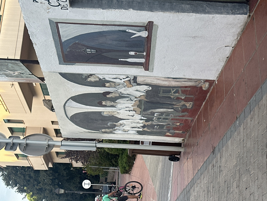 This image captures a street scene in Calella De Palafrugell, Spain, featuring a large mural on a building's white exterior wall.
The mural, centrally located, depicts an interior tavern scene. The left segment shows eight men: seven are seated around a square table, and one stands to the right of the table, appearing to serve. All men wear white shirts and dark trousers; several hold glasses or bottles. The background of this section features dark arched alcoves. The right segment of the mural portrays a painted window-like alcove containing a white wine bottle, a glass with red liquid, and a white, abstract vase or sculpture on a ledge. A dark, hanging lantern is depicted within this window scene.
The immediate foreground consists of a pedestrian sidewalk paved with reddish-brown square tiles directly adjacent to the mural wall, transitioning to grey interlocking pavers further left. A dark brown wheeled trash bin stands near a metal signpost on the reddish-brown tiled section.
To the left, a person wearing a green short-sleeved shirt, dark shorts, and a pink and white helmet is partially visible, walking or standing next to a black bicycle with pink accents on its frame and wheels. Behind them, an asphalt road is visible, flanked by a black street lamp and a white circular road sign with a horizontal bar. Green trees and shrubs line the background, with a multi-story, light-colored residential building featuring balconies and green window shutters visible beyond the foliage.
Above the mural on the right, partial text reading "CA" and "DE" is visible on the building wall. Overhead wires and a billboard with an abstract design are present in the upper background. The scene is illuminated by natural daylight.