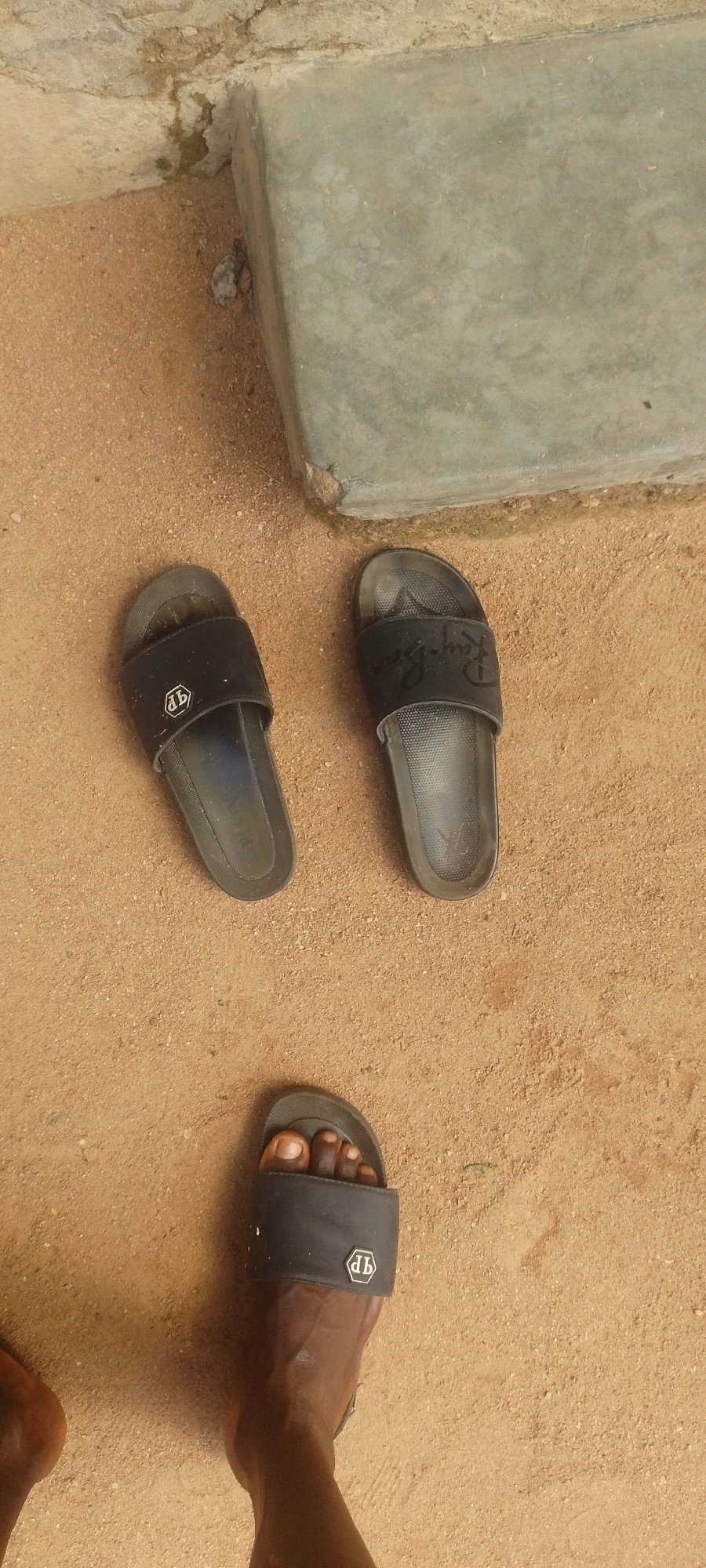 The media file shows a ground-level view of a sandy area with a concrete structure. Three black slides are visible. Two are placed side-by-side near the concrete structure, and a third slide is on the right foot of a person, partially obscuring their toes. The slides appear to be casual footwear, possibly for indoor or outdoor use. The sandy ground suggests an outdoor setting, and the presence of a concrete structure could indicate a pathway, wall, or foundation. The lighting suggests it is daytime. One slide has a visible logo that appears to be "Ray-Ban" written in script. Another slide has a hexagonal emblem with intertwined letters inside. The overall scene is simple and focuses on the footwear and the ground. The context of Jalingo, Nigeria, places the scene in a specific geographic location within West Africa.