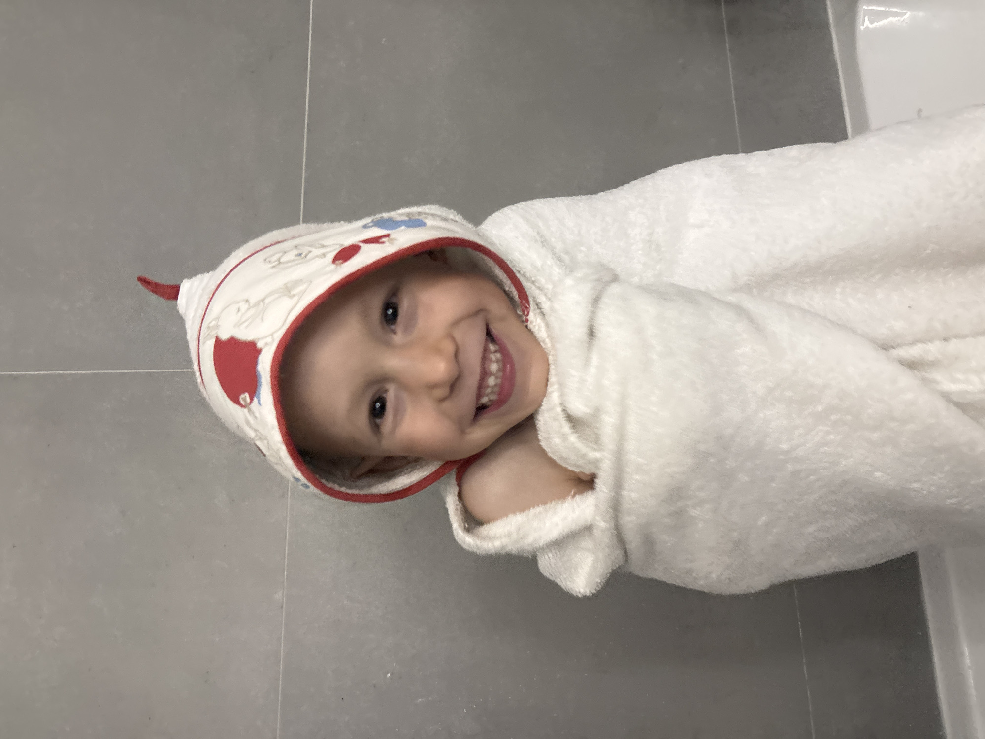 A young child, wrapped in a white hooded towel, lies on a gray tiled floor. The child is smiling and looking directly at the camera. The hood of the towel has a red trim and cartoon designs on it. The photograph appears to have been taken indoors, possibly in a bathroom, considering the tiled floor. There is no evidence suggesting that the photo was taken in the city of Chaikovsky, Russia.