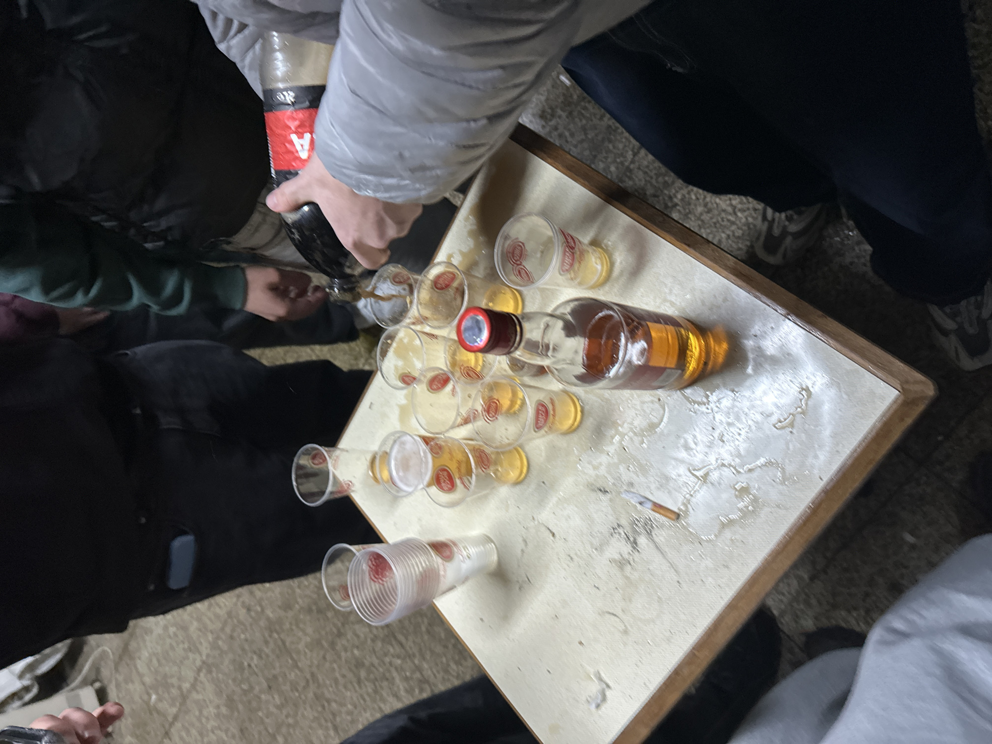 The scene depicts a group of people around a table in what could be a casual social gathering. A person is pouring a dark liquid from a bottle into glasses already containing an amber-colored liquid, presumably to make mixed drinks. There is also a bottle of the amber-colored liquor, a lighter, several empty plastic cups, and a lit cigarette on the table. The location is possibly in Bragança, Portugal, although there is no visual evidence to verify that.