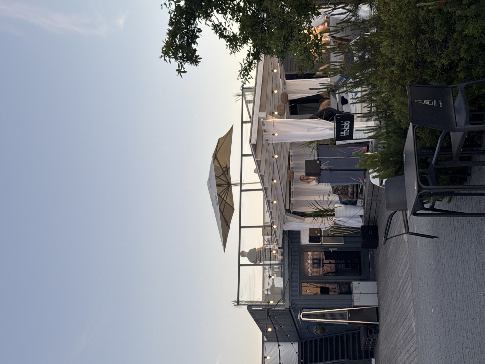 The image depicts an outdoor seating area with a contemporary, industrial-chic aesthetic, likely a cafe or bar, situated in Tallinn, Estonia. The scene is set in the late afternoon or early evening, with a clear sky and soft lighting.
Dominating the composition is a multi-level structure built from shipping containers, a common architectural trend in urban regeneration. The lower level appears to be the main establishment, with a dark facade, large glass doors, and windows revealing glimpses of interior lighting. A tall patio heater stands to the left of the entrance. Above this level, a second story extends, with a person standing near a glass railing, silhouetted against the sky.
The establishment features an open-air, covered dining or lounge area with white drapery, strings of fairy lights, and an open umbrella providing shade. Several potted plants, including tall green stalks in white pots, add a touch of nature to the urban setting. There are tables and chairs visible, some empty, suggesting a relaxed atmosphere. A DJ booth with speakers and equipment is set up in the foreground of this area, with a woman standing behind it. A sign displaying "fritz-kola" is prominent.
The foreground shows a paved area with a grey brick pattern and a wooden deck. Two dark-colored outdoor tables with chairs are positioned for patrons. Beyond the immediate seating area, lush green bushes and trees are visible, softening the industrial elements. The overall impression is a trendy, urban spot designed for outdoor socializing and entertainment. The specific location in Tallinn is not explicitly stated in the visual evidence, but the architectural style and atmosphere are consistent with modern urban leisure spaces.