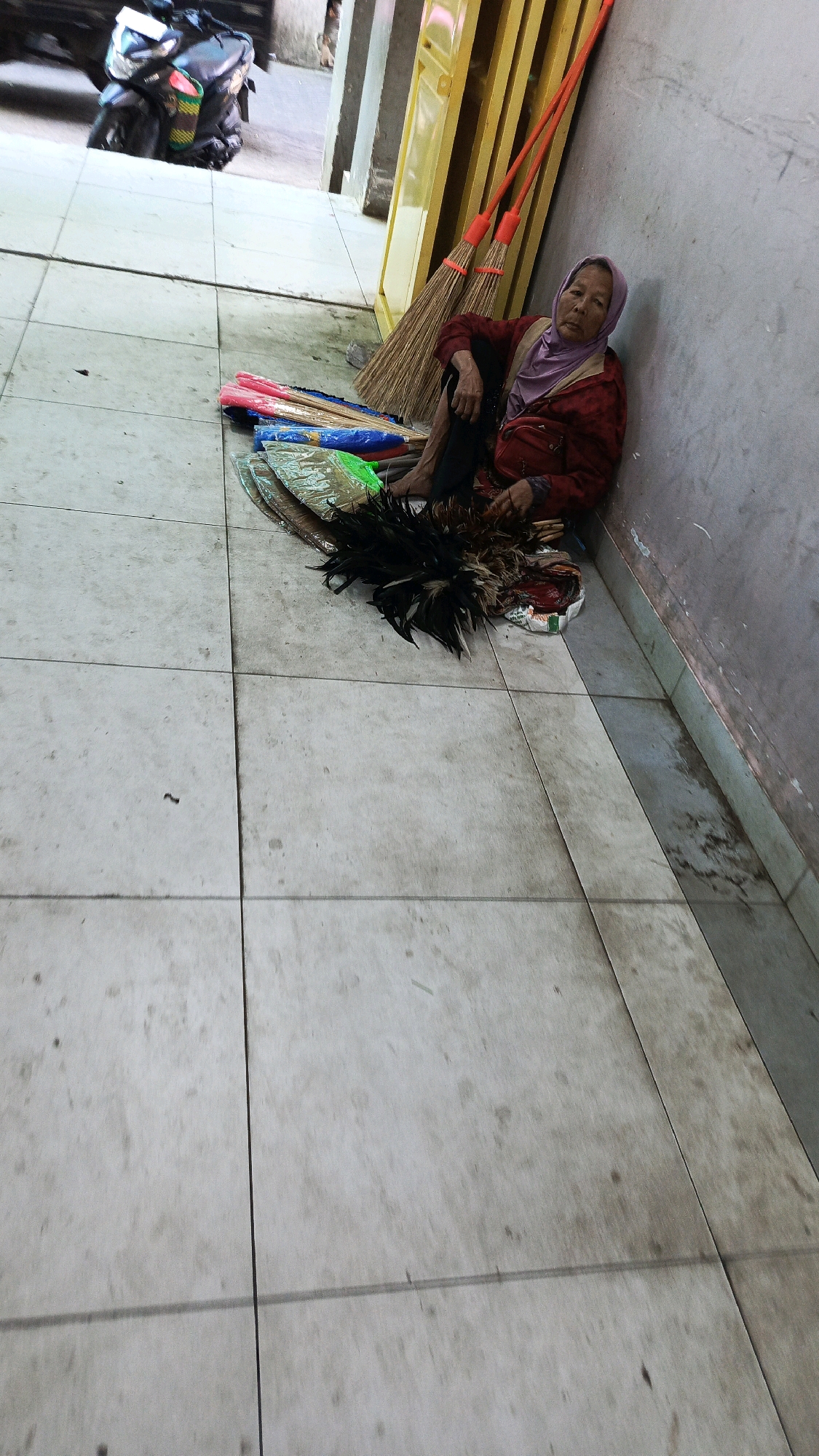 An elderly woman, wearing a purple headscarf and a red jacket over a purple top, sits on a tiled floor next to a light gray wall. She is leaning against the wall and appears to be resting or waiting. In front of her are several items for sale, including brooms with orange handles and bristles of various colors, some wrapped in plastic, and a pile of dark feathers. Two long brooms with orange handles are propped against a bright yellow structure to her right. The floor is tiled in a light neutral color and shows some signs of dirt and wear. To the left, a dark motorcycle with colorful bags attached is partially visible. The setting appears to be an outdoor or semi-outdoor area, possibly a market or a walkway in Ponorogo, Indonesia, given the architectural elements and the presence of items typically sold in such locations.