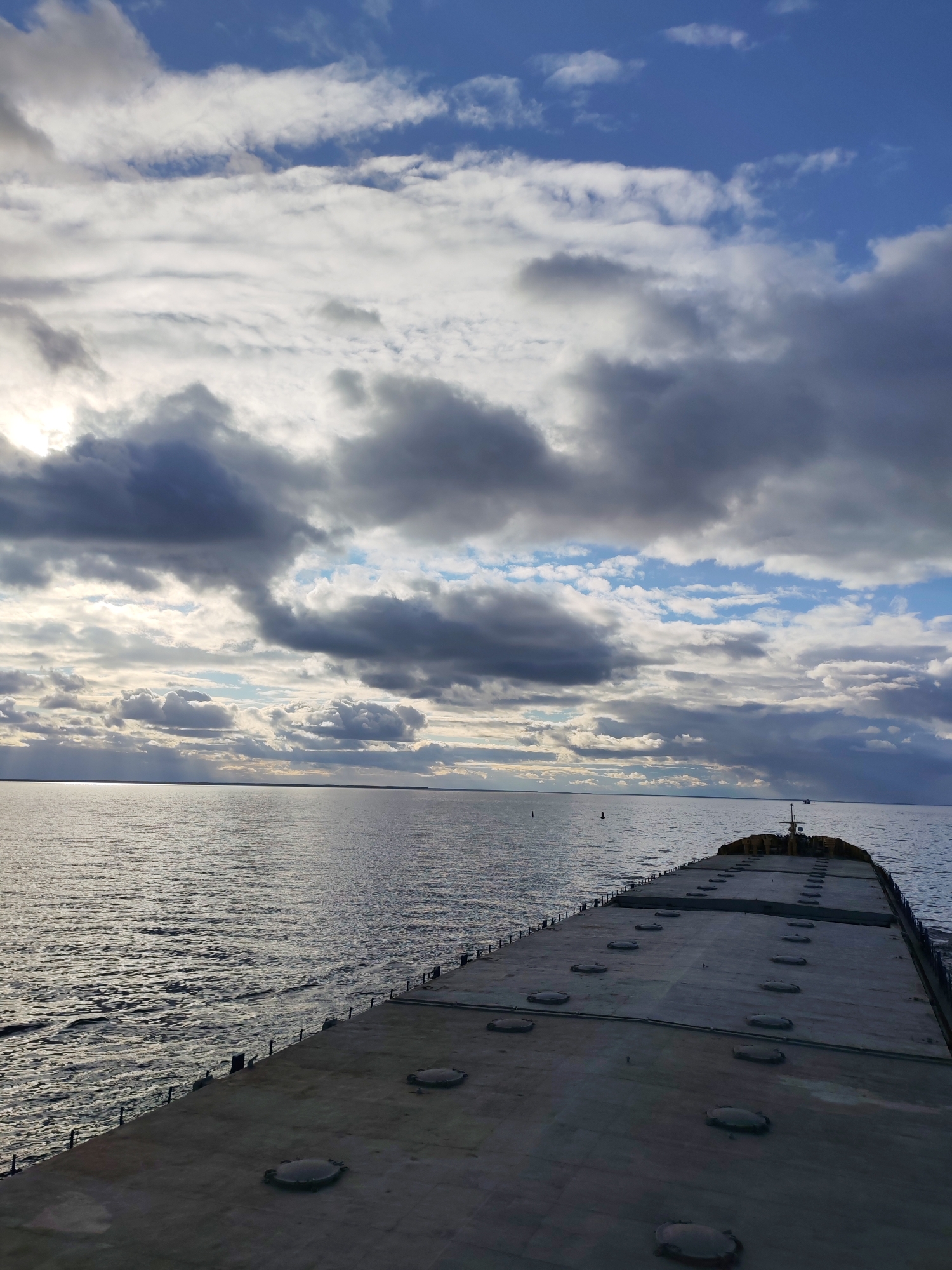 The image displays a wide-angle view from the stern of a large, flat-deck cargo vessel, likely a barge or bulk carrier, traversing a vast body of water. The deck, painted in a dark gray, occupies the lower portion of the frame and features numerous circular hatch covers arranged in rows, receding towards the distant bow. A railing with stanchions is visible along the starboard side of the vessel, where the water churns slightly.
The expansive body of water, which appears to be a large river or reservoir based on the provided location context of Novaya Svobodka, Russia, stretches to a low horizon. The water surface shows gentle ripples and reflects the overcast sky. In the mid-distance on the water, two buoys are visible, indicating a marked waterway.
The sky is dramatic and dynamic, filled with a mix of heavy, textured gray and white clouds that partially obscure the sun. Patches of brighter blue sky are visible in the upper right. On the left horizon, sunlight breaks through the clouds, creating a bright glare on the water and subtle crepuscular rays. The lighting suggests either late afternoon or early morning. No people or visible text are present in the image. The scene evokes a sense of journey and the vastness of the natural environment under a varied sky.